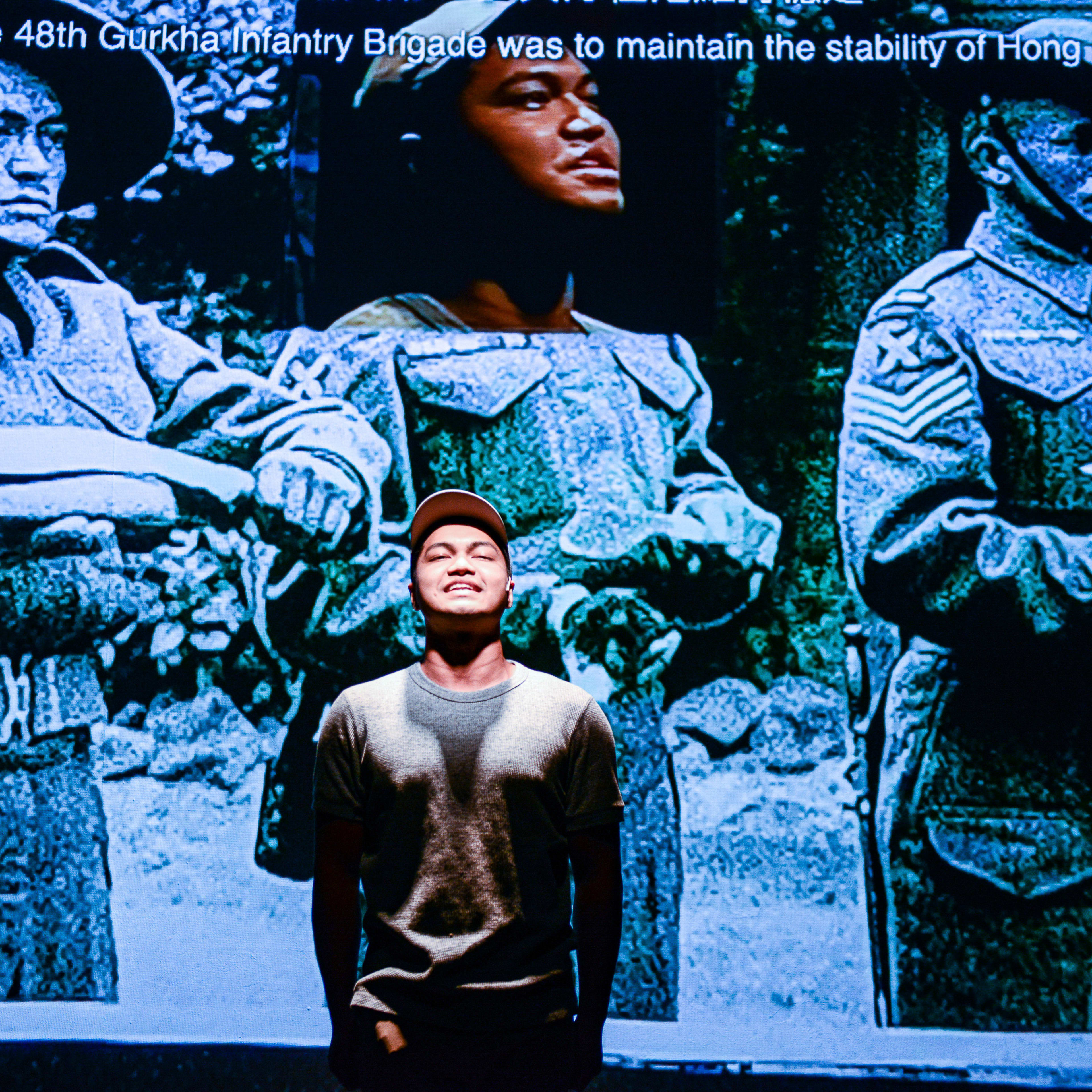 在天台製作（Rooftop Productions）的當代劇場作品《山下的證詞》（Testimony）中，演員 Basnet Tusar 站在舞台中央，被一束聚光燈照亮。他身後的巨型投影展示了一張泛藍的黑白歷史照片，呈現了啹喀兵在香港的軍事歷史背景。在演出中，他語帶深意地說道：「你說一切都會被毀掉？那為什麼要戰鬥？」