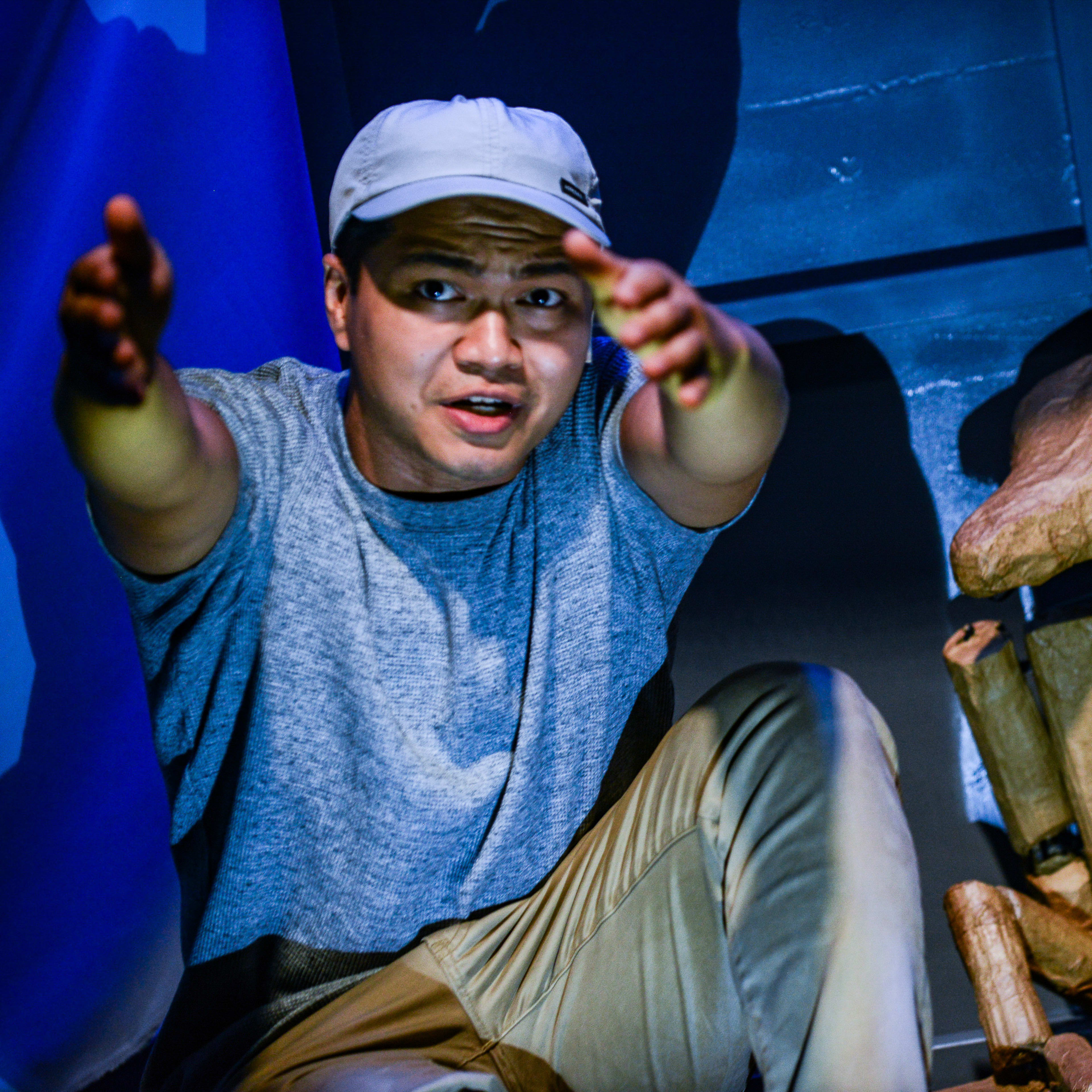 天台製作（Rooftop Productions）當代劇場作品《山下的證詞》演出劇照。演員 Basnet Tusar 在藍色調的舞台燈光下，神情專注且急切地向鏡頭伸手，身旁坐著一個以紙板製成的等身偶，隱沒在白紗幕後。演出中他説道：「我們會整晚在整條中港邊界留守。」