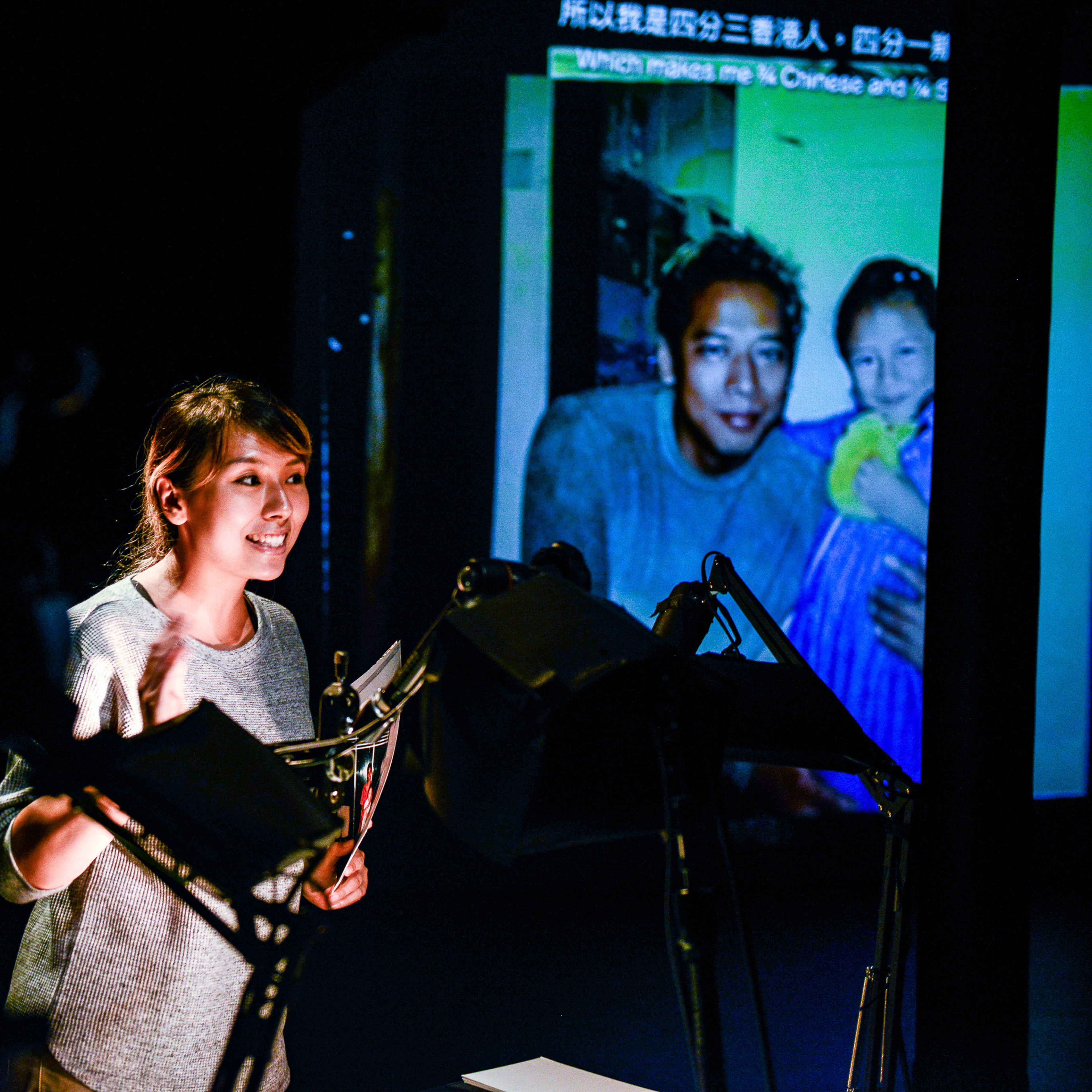 天台製作（Rooftop Productions）的當代劇場作品《山下的證詞》（Testimony）現場演出。在充滿戲劇張力的燈光下，演員李里莎（Arisa Yasaratne）於昏暗的舞台上進行獨白演出，背景投影著一張模糊的家庭合照，畫面中是一名男子與一名小女孩。此演出以獨特的視覺敘事手法探討身分認同與個人經歷。李里莎感性地訴說：「我媽媽是百分百的華人，在香港出生，我爸爸是半斯里蘭卡人。」