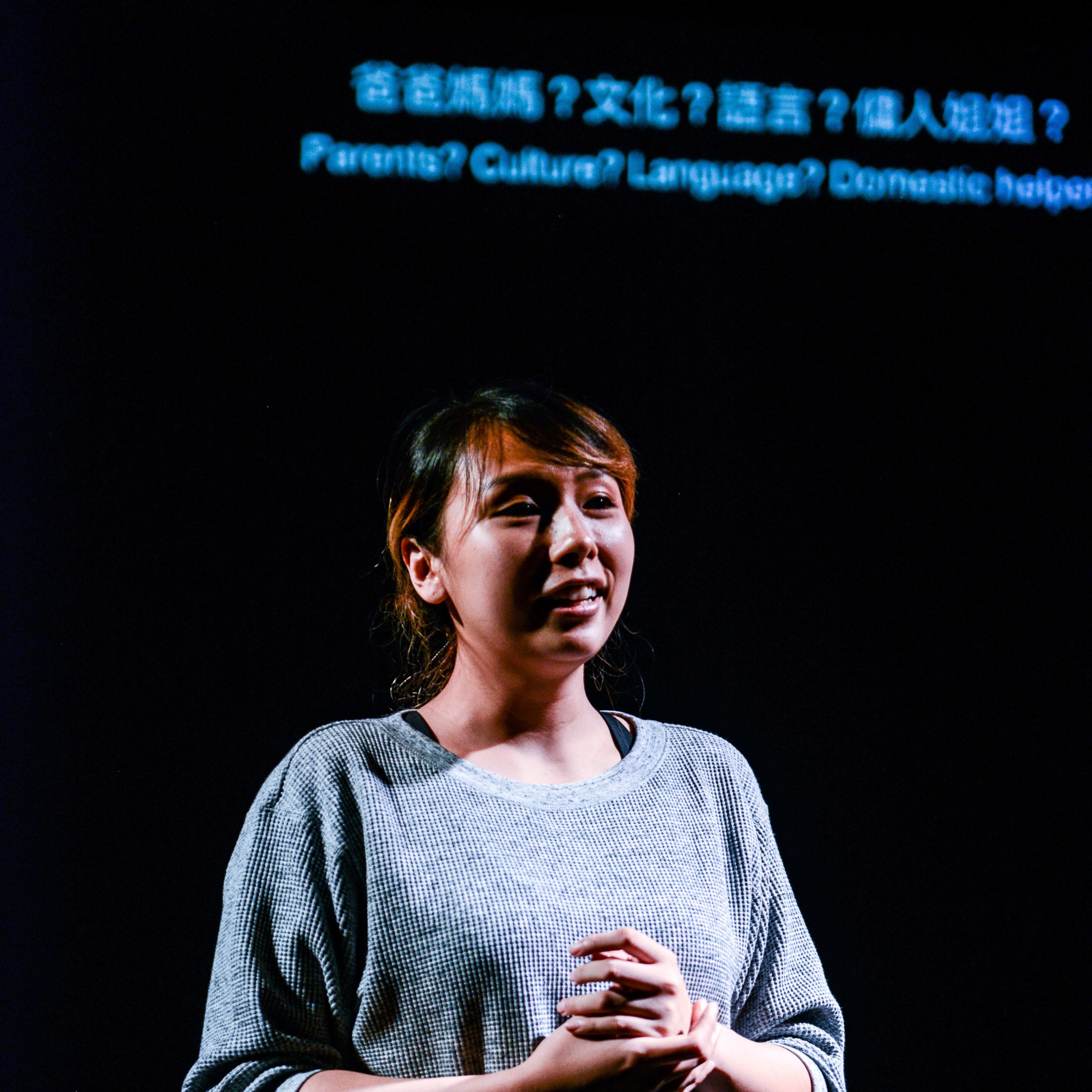 在天台製作的當代香港劇場作品《山下的證詞》（Testimony）中，演員李里莎站在舞台中央，身後是一片漆黑，聚光燈打在她的身上。她身穿灰色質感上衣和卡其色褲子，雙手交疊於胸前，神情專注而感性地與觀眾對話。她頭頂方的投影屏幕上顯示著：「爸爸媽媽？文化？語言？傭人姐姐？」。 「《山下的證詞》」