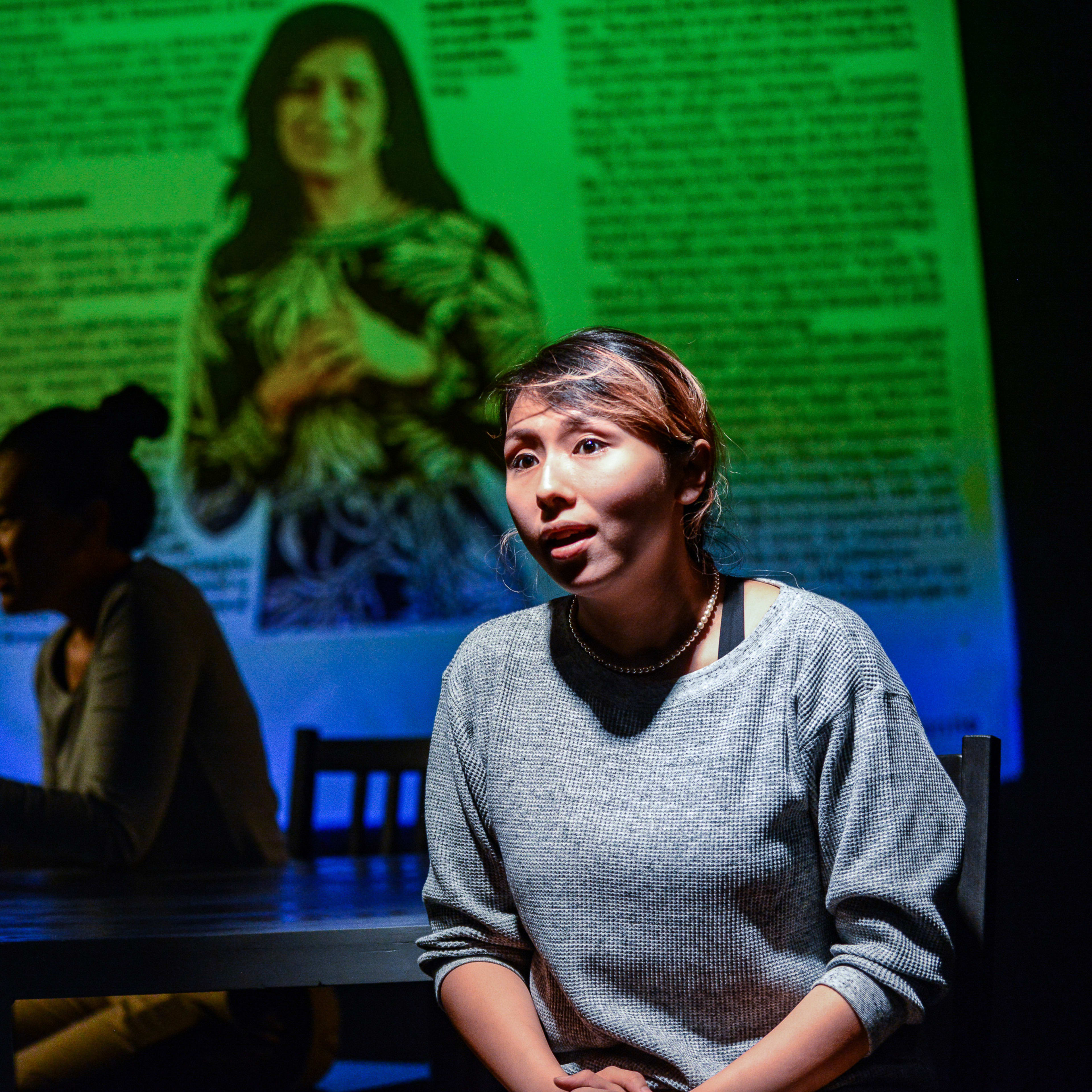 在天台製作的當代劇場作品《山下的證詞》（Testimony）中，演員李里莎（Arisa Yasaratne）坐在舞台前方，在冷色調的聚光燈下進行獨白。背景投射著一張泛著綠光的女性肖像及文獻資料，營造出強烈的紀實感與敘事氛圍。舞台後方另一位演員正坐在桌旁，演繹著這場充滿戲劇張力的演出。李里莎在戲中回憶道：「1993年，我在羅兵咸永道會計師事務所開始工作。」