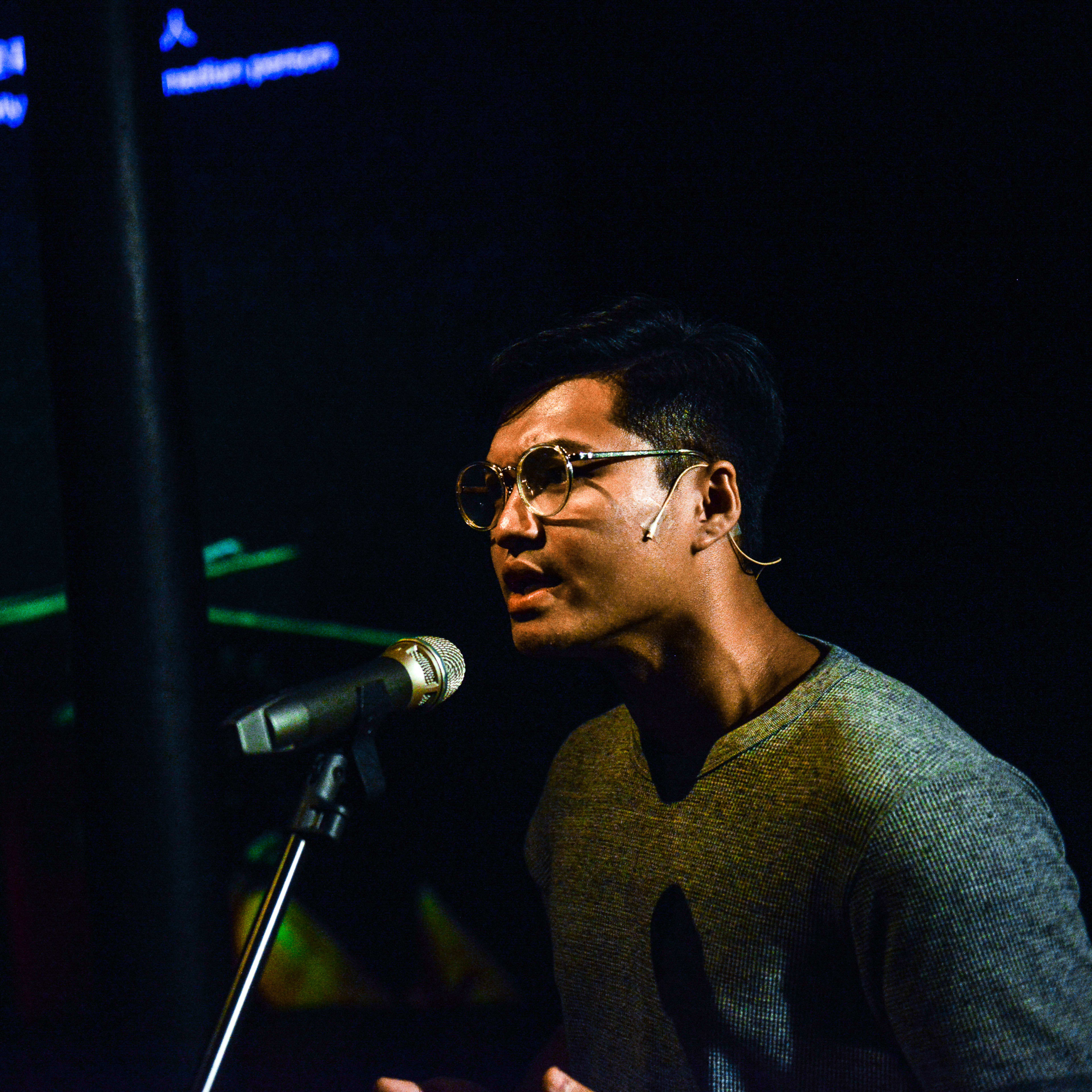 在天台製作（Rooftop Productions）的當代劇場作品《山下的證詞》（Testimony）中，演員 Basnet Tusar 在低調的舞台燈光下神情專注地對著麥克風進行演出。背景投影著護照的綠色光影，營造出強烈的戲劇氛圍。《山下的證詞》