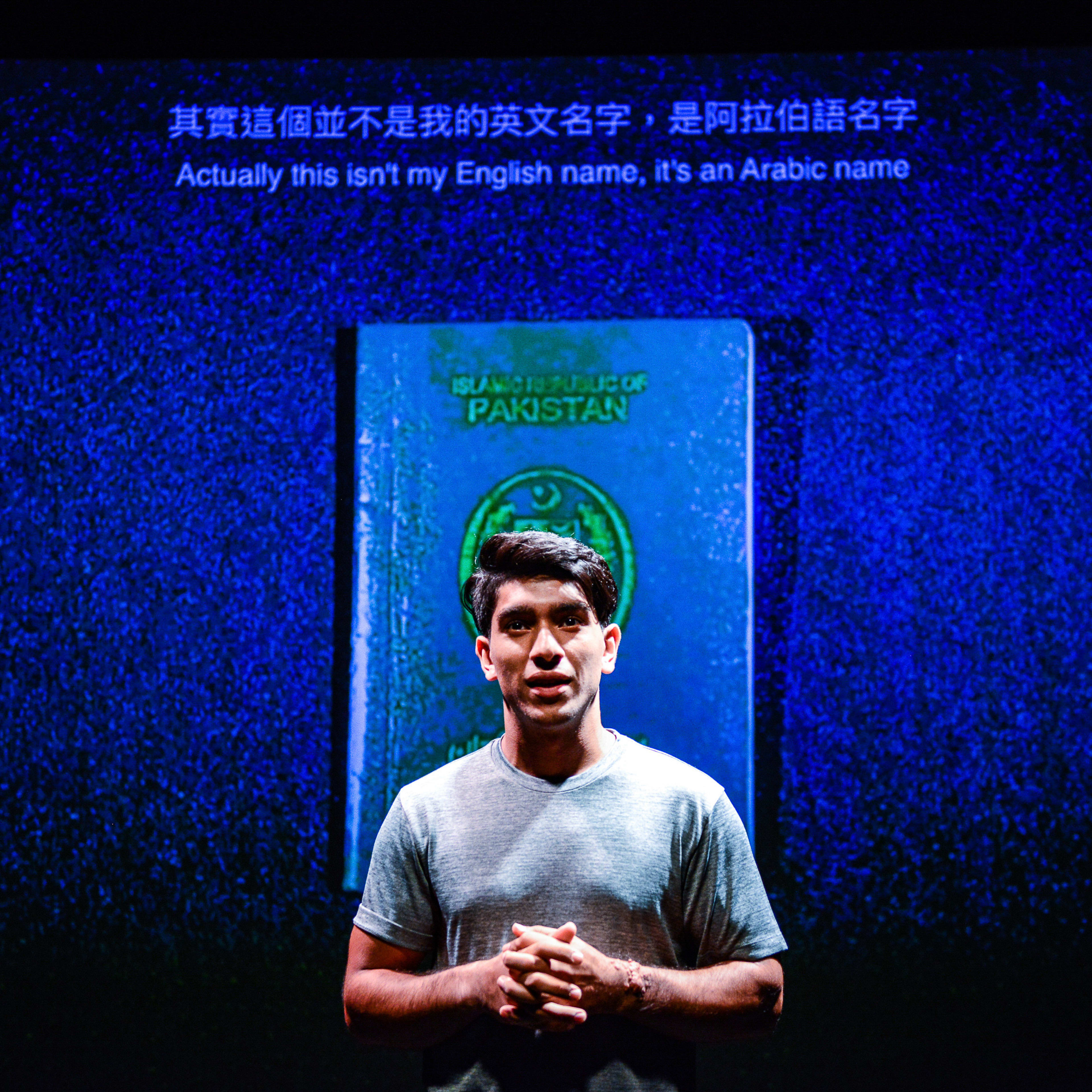 天台製作當代劇場作品《山下的證詞》（Testimony）的舞台劇照。演員巫加沙（Mohammad Kashif Ali）站在舞台中央，在深藍色背景的襯托下，身後投影出巨大的巴基斯坦護照影像。他神情真摯、語氣平靜地與觀眾交流。劇中演員講述道：「其實這個並不是我的英文名字，是阿拉伯語名字。」