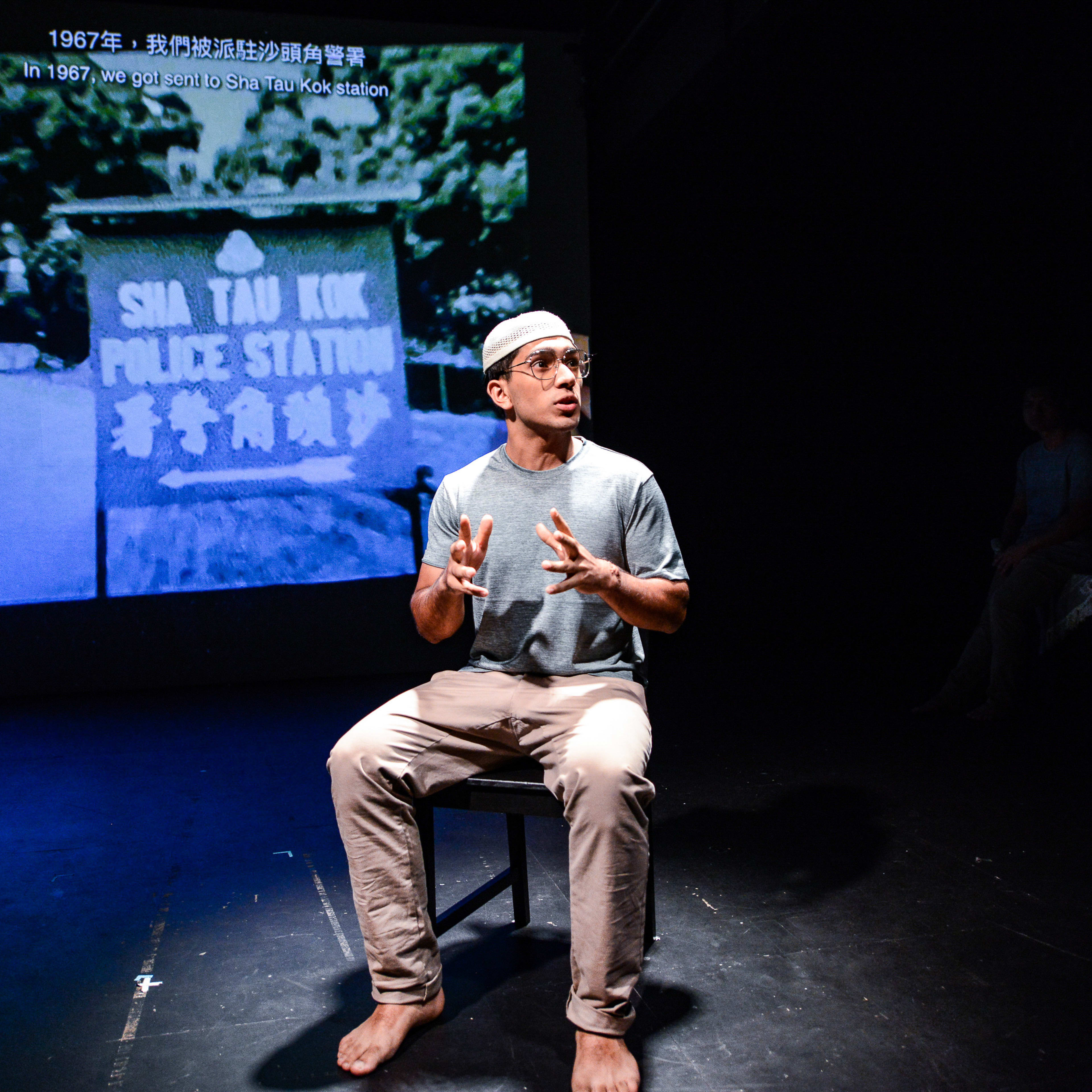 在天台製作（Rooftop Productions）當代劇場作品《山下的證詞》（Testimony）的演出劇照中，演員巫加沙（Mohammad Kashif Ali）坐在舞台中央的一張木椅上，身處幽暗極簡的劇場空間，僅有一束強光聚焦於他。他身穿灰色T恤與淺色長褲，正以生動的手勢講述劇中的歷史證詞。背景投影出一張高對比度的黑白照片，畫面中可見寫有「沙頭角警署」的指示牌，並標註著1967年的歷史背景。演員在演出中感嘆道：「然後有1000人來到沙頭角邨，沿著在邊界大叫。」