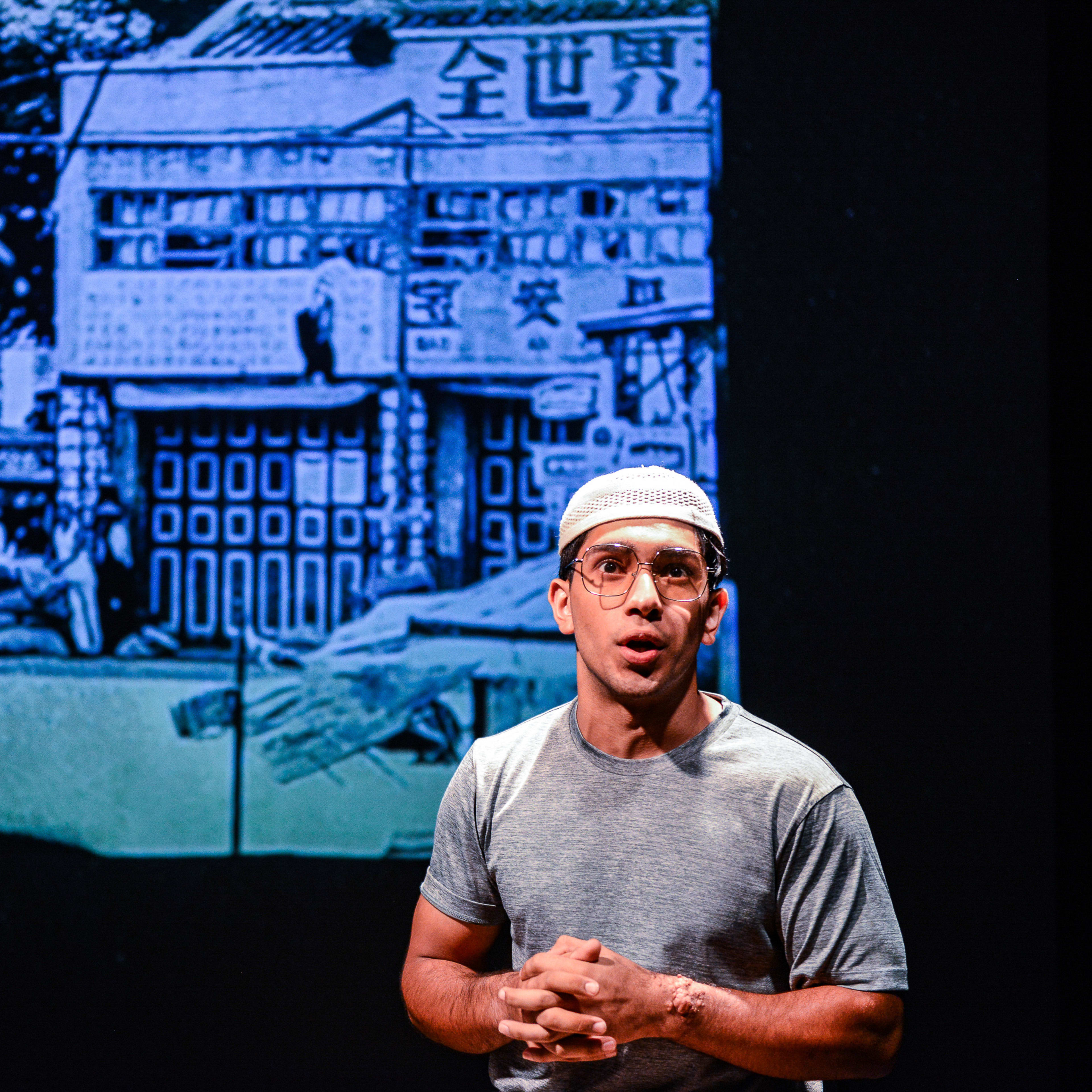 天台製作（Rooftop Productions）當代劇場作品《山下的證詞》（Testimony）的劇照。演員巫加沙身穿簡約灰色T恤，頭戴白色小帽，站在舞台上，神情專注而感性，正向觀眾深情訴說。他身後的巨型投影呈現出一幅帶有藍色調的香港舊街道景象，畫面中隱約可見傳統建築與中文招牌，在表演者與背景之間形成了強烈的視覺張力。《山下的證詞》