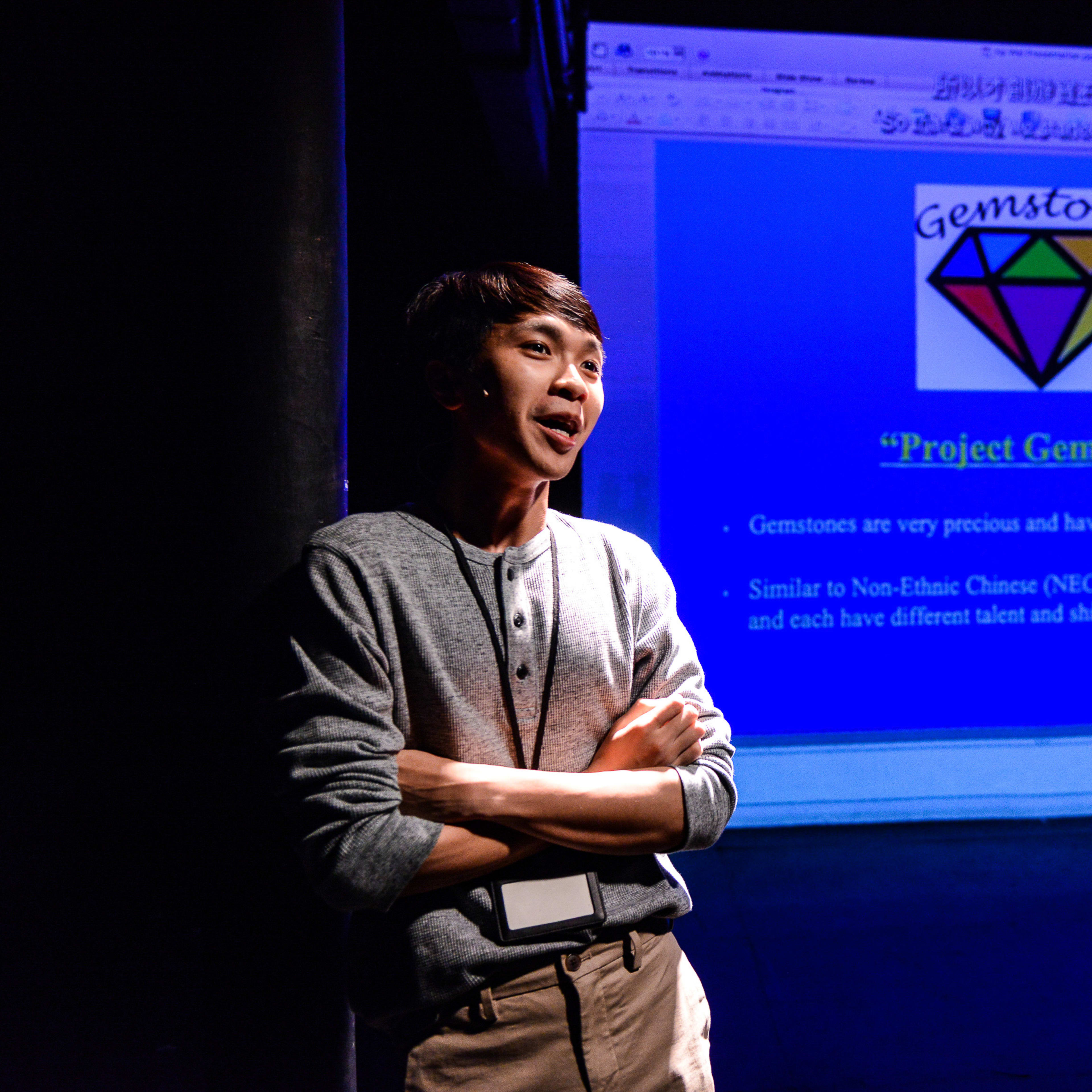 在天台製作（Rooftop Productions）的戲劇作品《山下的證詞》（Testimony）中，演員吳景隆站在舞台上，雙臂交叉，在黑暗的背景中被聚光燈照亮。他身後的投影幕顯示著名為「Project Gemstone」的演示文稿，圖中印有一個彩色鑽石圖案，文字將寶石的價值與非華裔（NEC）學生的多元才華作出了比喻。《山下的證詞》