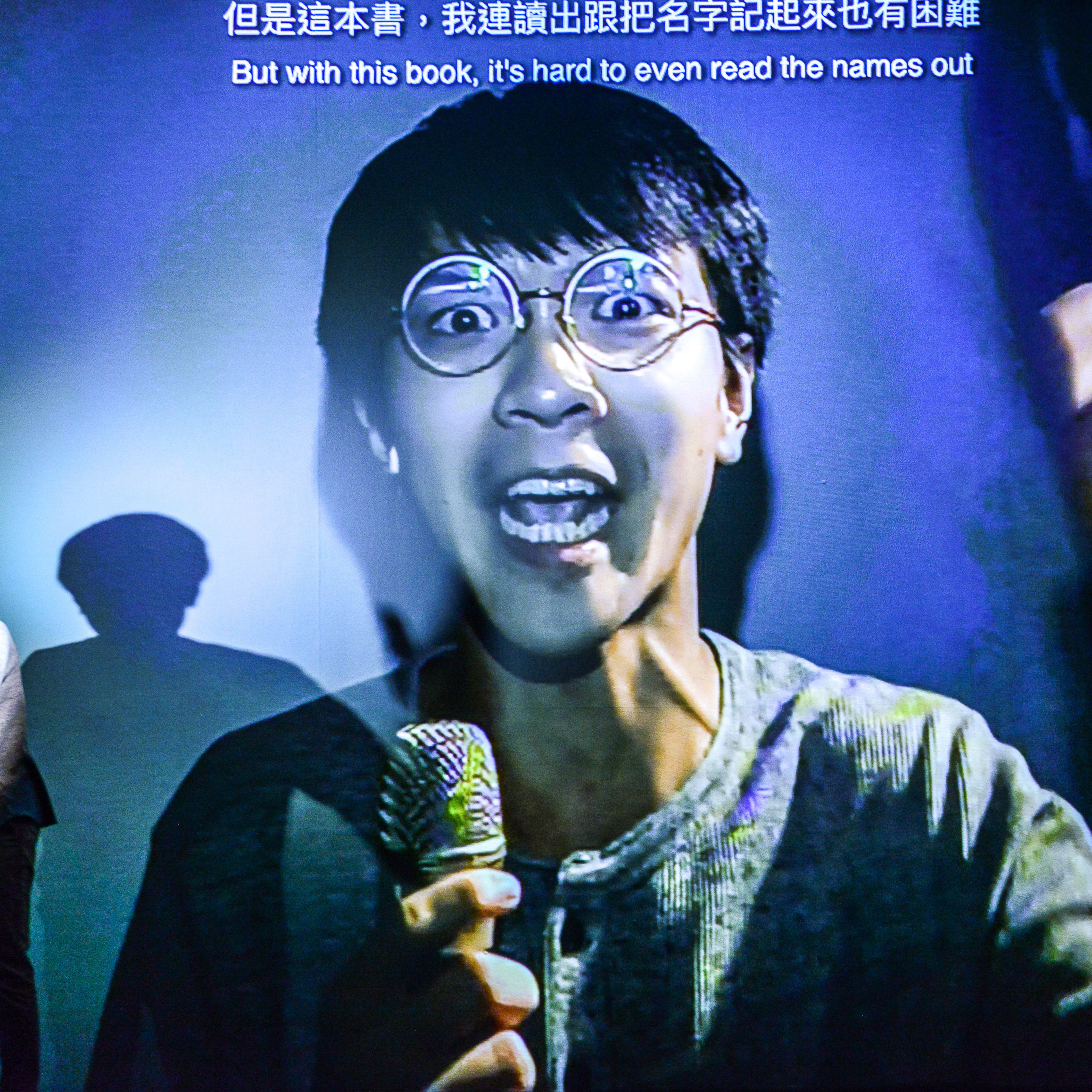 在天台製作（Rooftop Productions）的戲劇演出《山下的證詞》（Testimony）中，演員吳景隆站在昏暗的舞台上，身後投射出冷色調的藍色影像。投影畫面中，戴著圓框眼鏡的吳景隆表情生動地對著麥克風演繹，與舞台上真實的演員形成虛實交錯的視覺效果。劇場燈光突顯了現場表演與多媒體影像的互動張力。吳景隆在表演中說道：「雅典人成立的民主制度、文藝復興、秦始皇。但是這本書，我連讀出跟把名字記起來也有困難。」