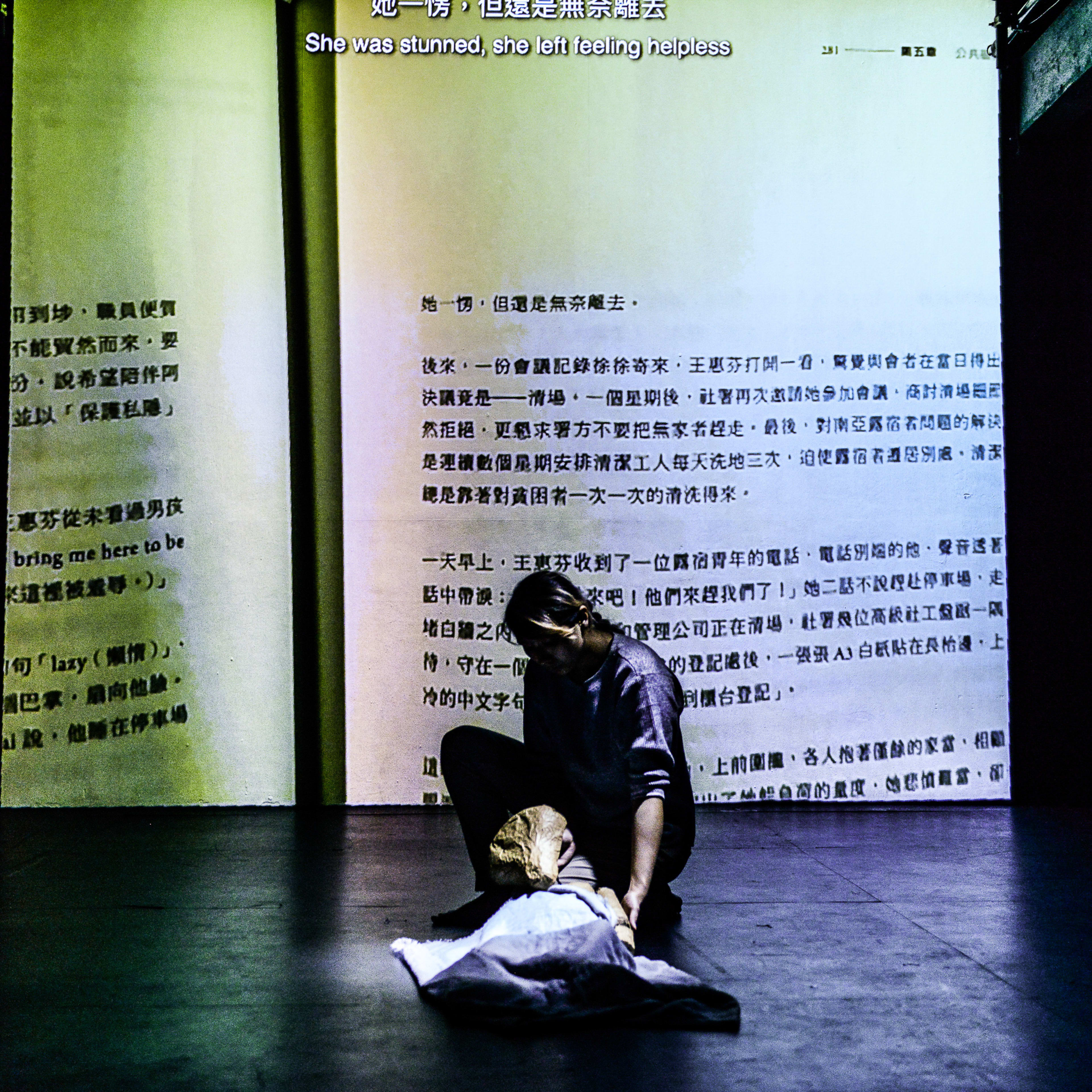 在天台製作 (Rooftop Productions) 的當代劇場作品《山下的證詞》(Testimony) 中，一位演員於昏暗的舞台上跪地，身旁覆蓋著一具用布遮蓋的形體，背景投映著大幅的文獻文字，呈現出強烈的歷史沉重感與當代劇場的敘事張力。這一幕展現了演出中深刻的情感張力與舞台美學。劇中有一句對白：「牆上面，用紅色的筆寫著『沒有將來』。」