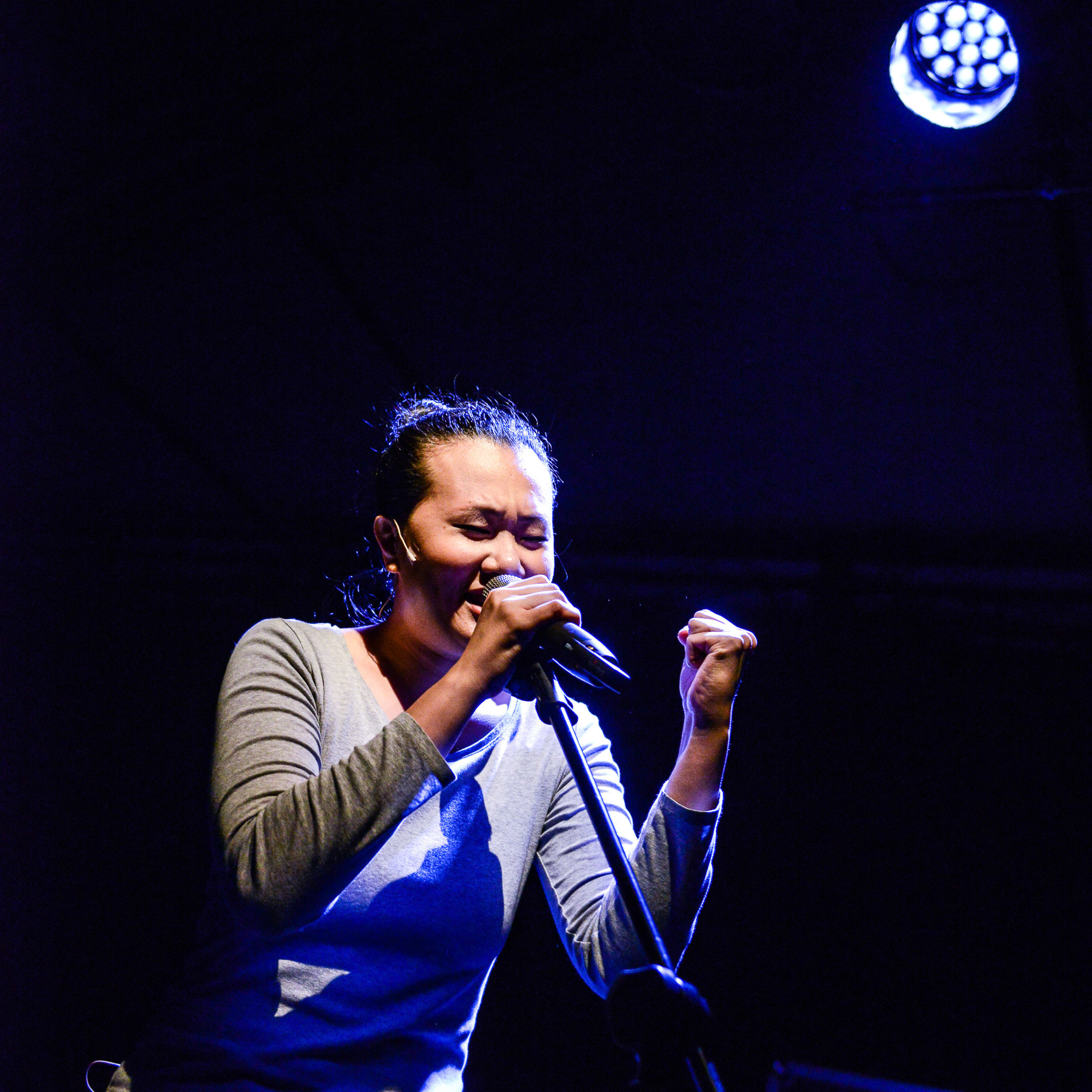 演員李婉晶在天台製作的當代劇場作品《山下的證詞》（Testimony）演出中，充滿感情地對著麥克風歌唱。在戲劇性的藍色舞台燈光下，她緊握左拳，神情投入，展現了演出中極具張力的一幕。「《山下的證詞》」