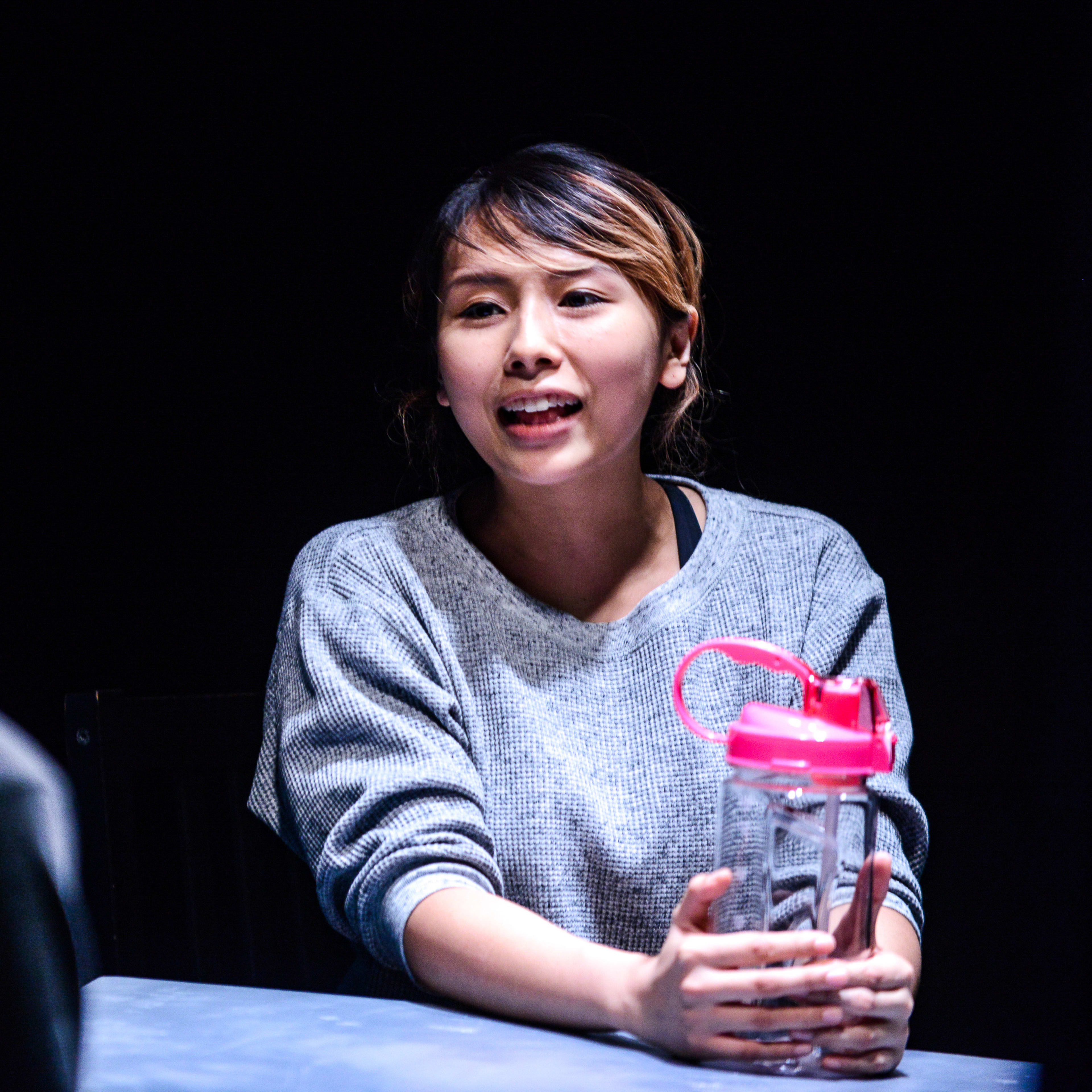 天台製作的當代香港劇場作品《山下的證詞》（Testimony）中，演員李里莎（Arisa Yasaratne）在漆黑的舞台背景下，被聚光燈照亮，手持水瓶神情專注地進行演出。「但是我身邊大部分的同學，也是在讀本地小一的中文。」