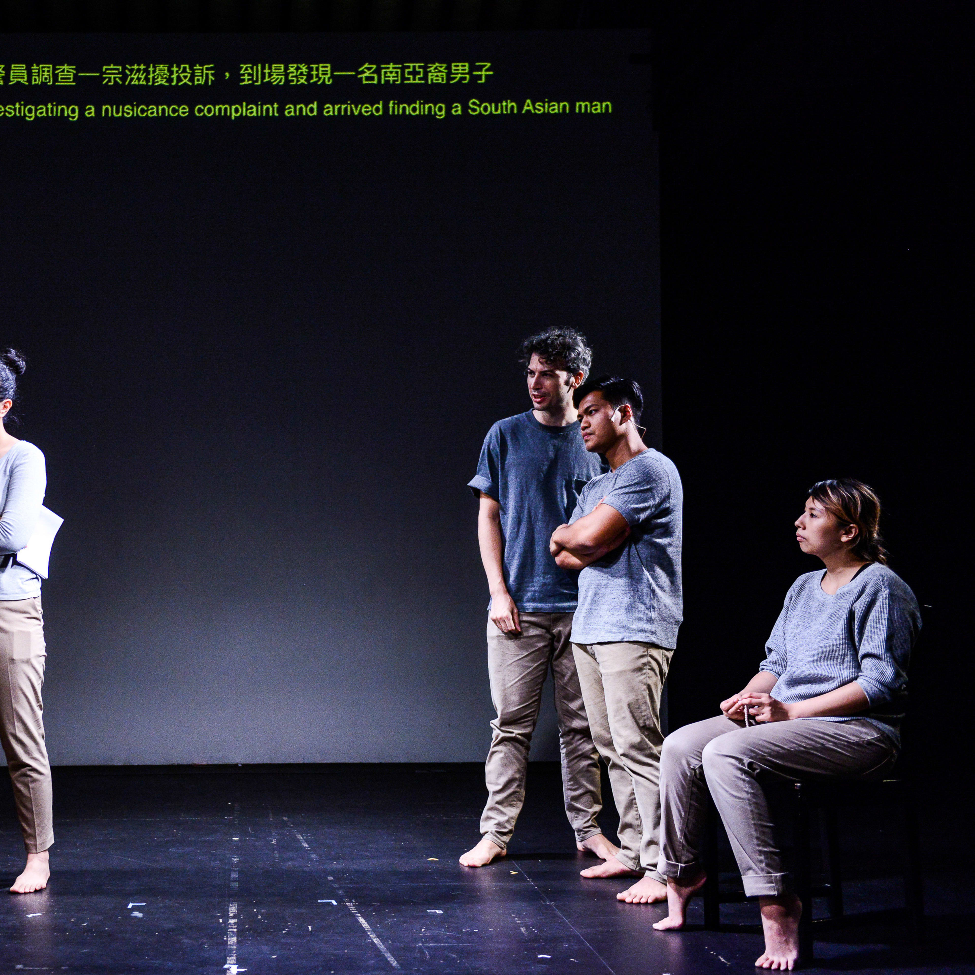 天台製作（Rooftop Productions）當代劇場作品《山下的證詞》（Testimony）的演出劇照。五位演員站在簡約的黑色舞台上，舞台後方的投影幕顯示出關於警員處理南亞裔男子滋擾投訴的文字。透過極具張力的肢體劇場，該劇深入探討香港複雜的社會議題。現場演員發出詰問：「Tusar，你知道林寶案嗎？」