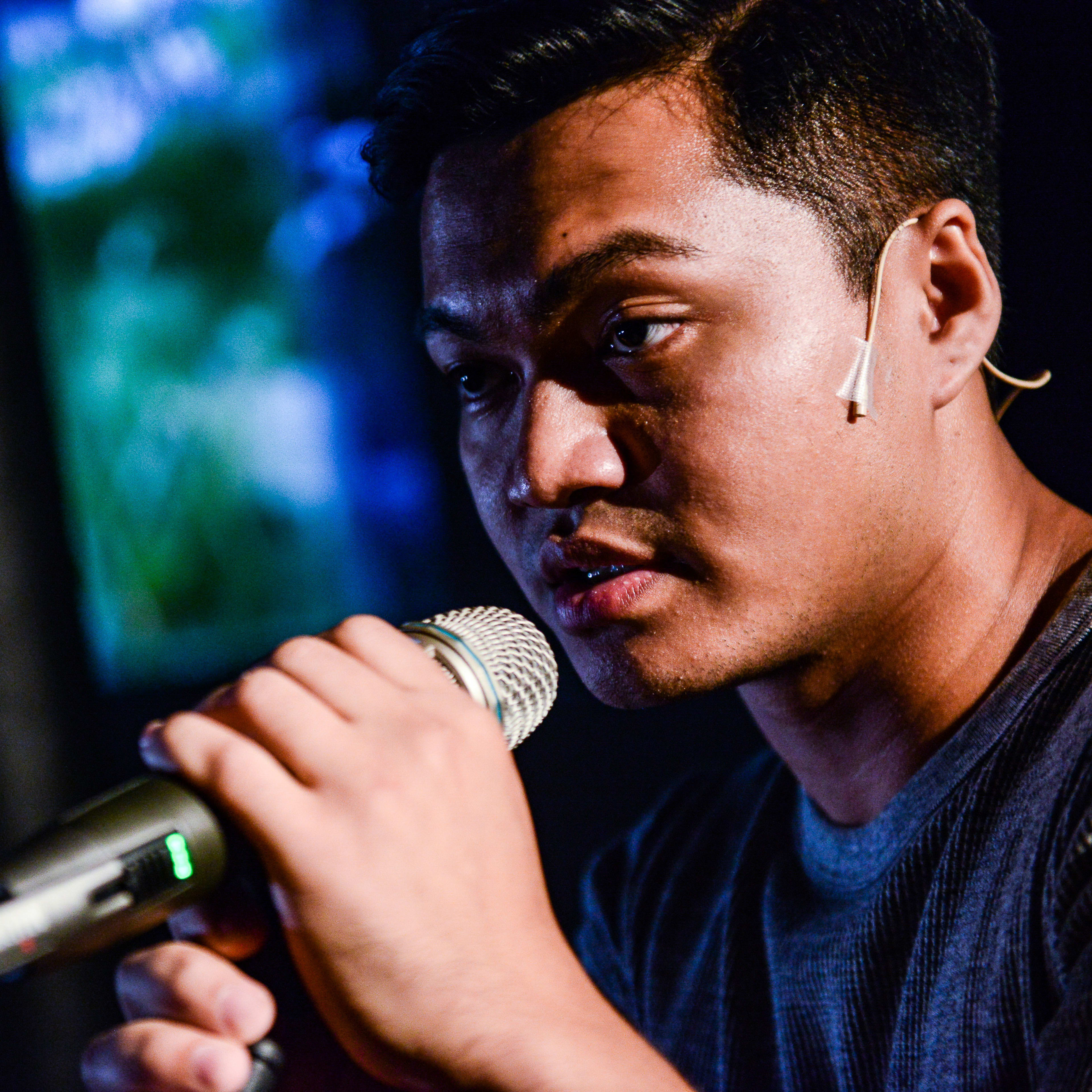 演員 Basnet Tusar 於天台製作（Rooftop Productions）當代香港劇場作品《山下的證詞》演出期間，在充滿戲劇感的藍色燈光背景下，神情凝重地手持麥克風發言。這張特寫鏡頭捕捉了角色訴說劇中台詞的動人瞬間：「造就你嗅不到的景氣」。
