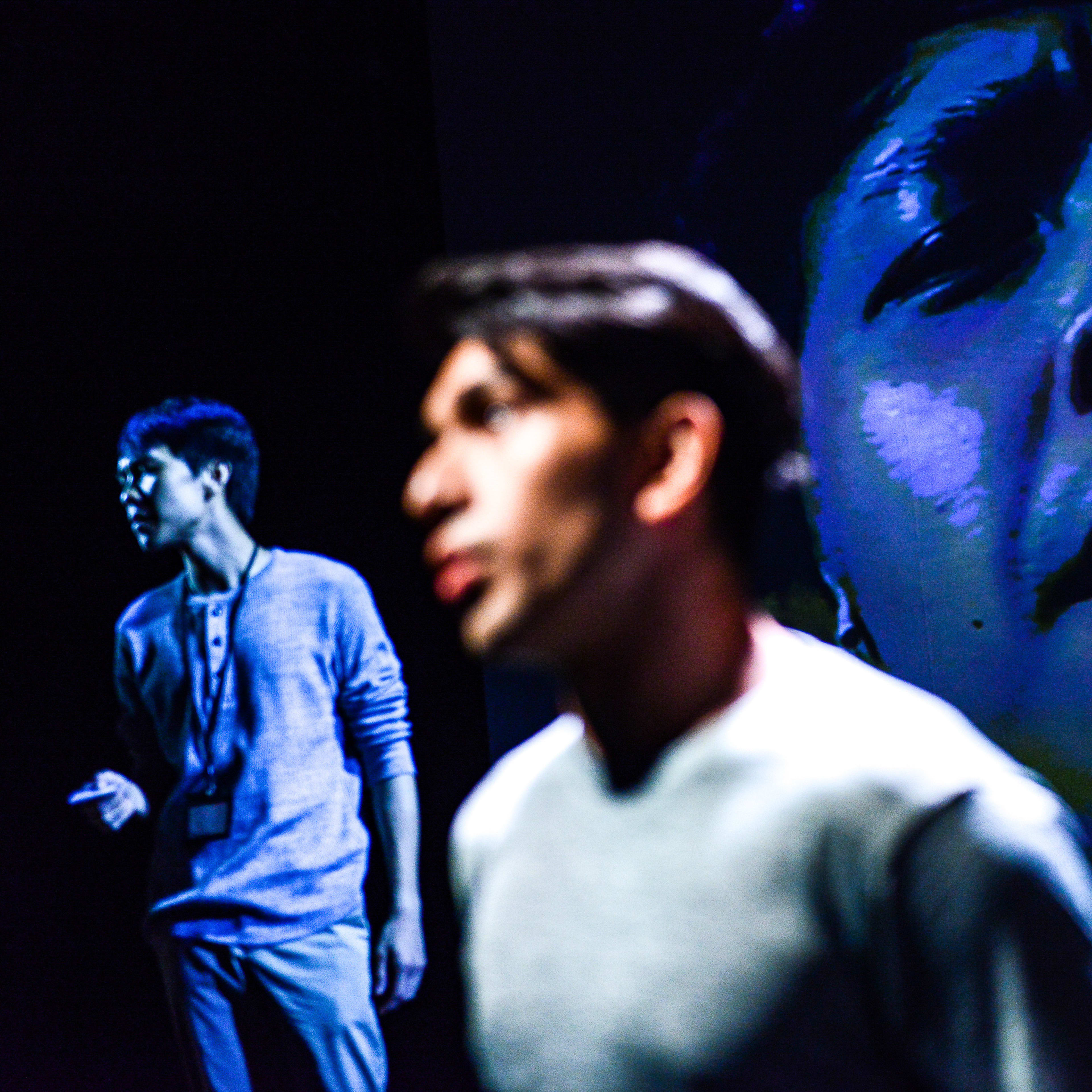 在天台製作（Rooftop Productions）的當代劇場演出《山下的證詞》中，演員巫加沙處於前景，神情深刻而凝重。舞台後方，另一位演員在暗處隱約可見，而背景投影著一張巨大、高對比度的藍色調人臉，為這場香港劇場演出增添了震撼的視覺層次感。巫加沙唸道：「我第一次被查身份證的時候，我在屋企樓下」