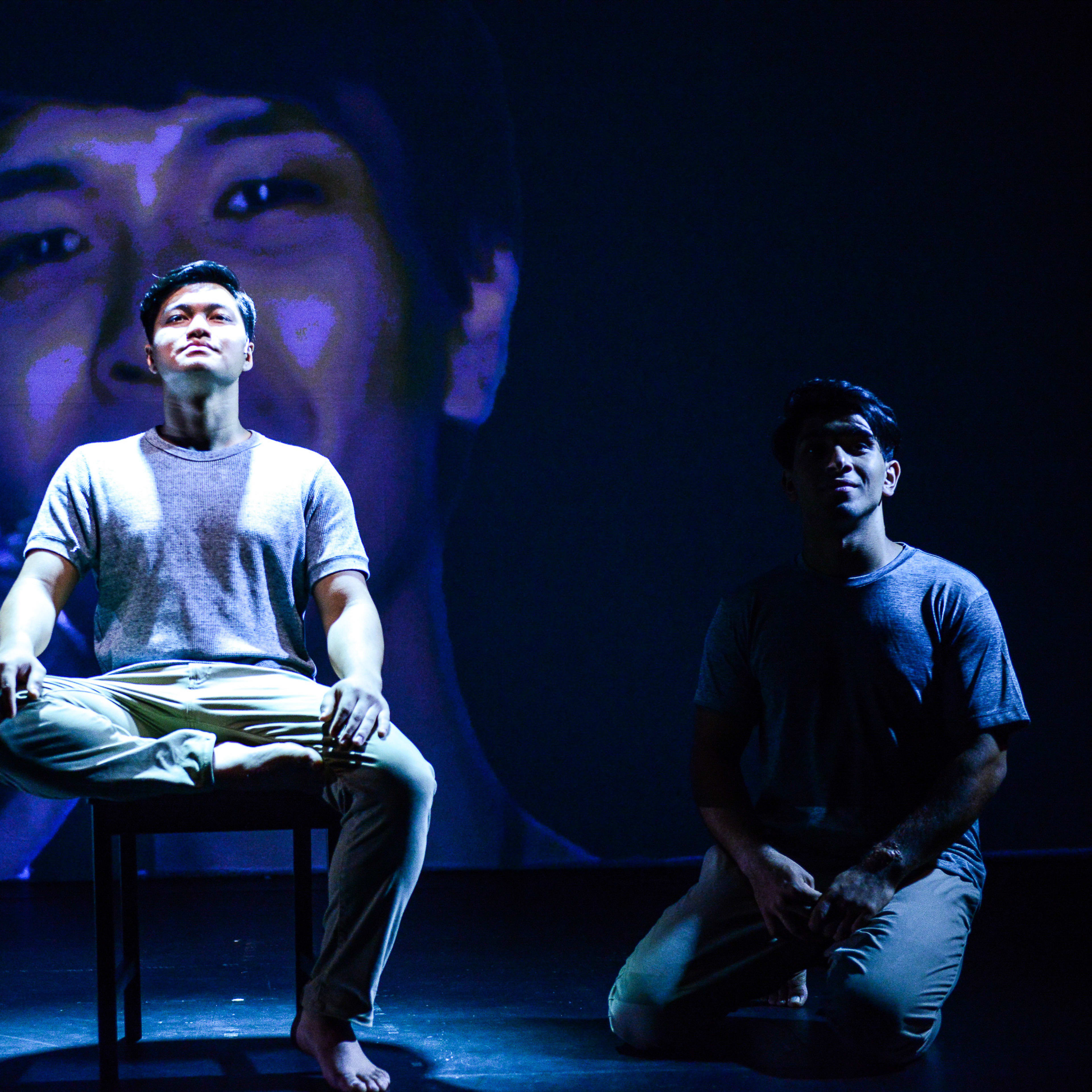 在天台製作（Rooftop Productions）的當代劇場作品《山下的證詞》（Testimony）中，一名演員在舞台中央的椅子上盤腿而坐，沐浴在強烈的藍色聚光燈下。他身後巨大的投影映出他模糊的臉龐，呼應著沉思的神情。舞台右側，另一位演員跪在陰影中，同樣被冷調的藍光籠罩，為這場香港劇場演出營造出充滿戲劇張力的氛圍。《山下的證詞》