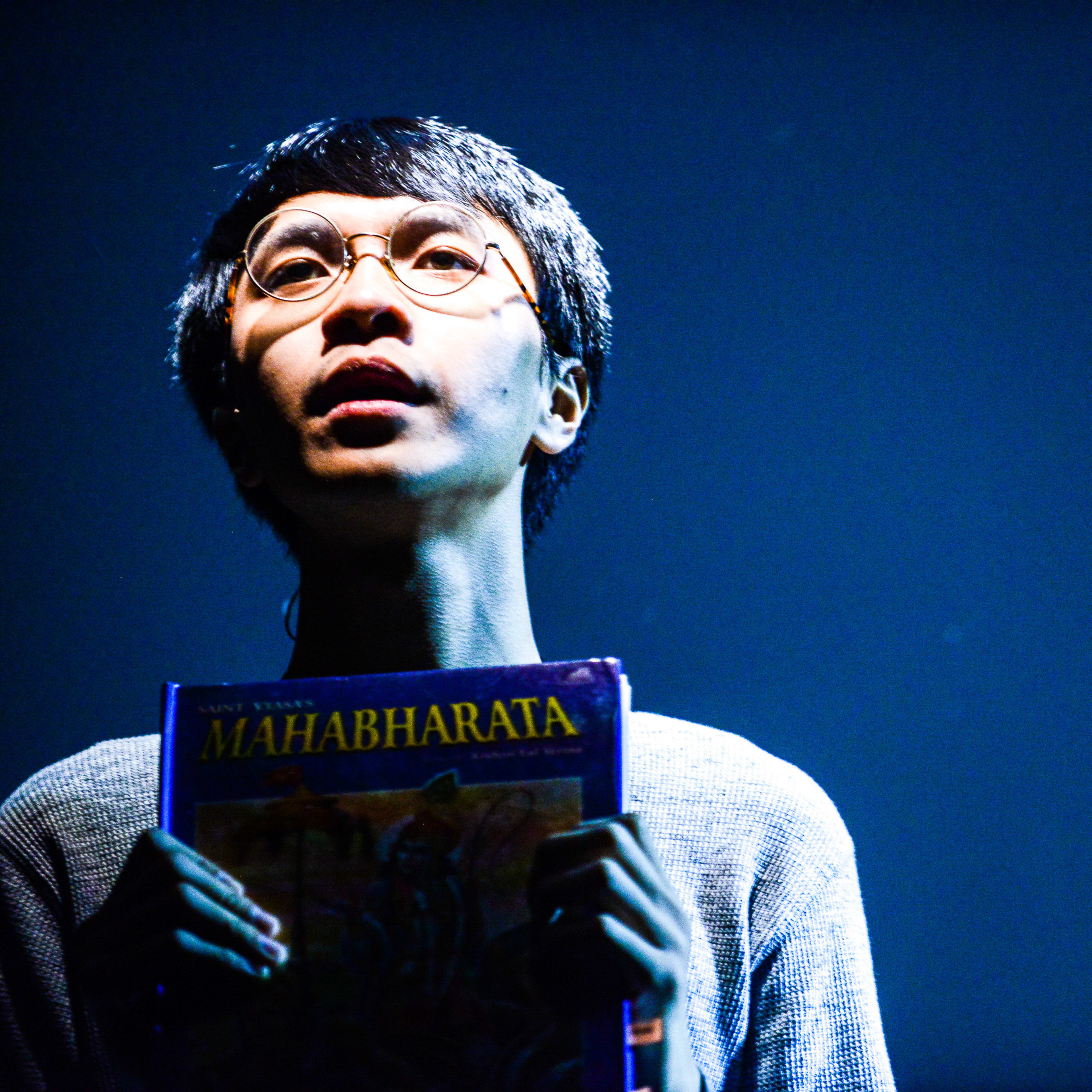 在天台製作（Rooftop Productions）當代香港劇場作品《山下的證詞》（Testimony）的一幕中，演員吳景隆在冷調藍光的舞台上，雙手捧著一本《摩訶婆羅多》（Mahabharata）。他神情專注地望向遠方，流露出深沉的思考與省察。在演繹這段關於歷史記憶的劇目時，他緩緩說道：「將來並不存在，但我會給你一雙眼睛去看一看戰爭」