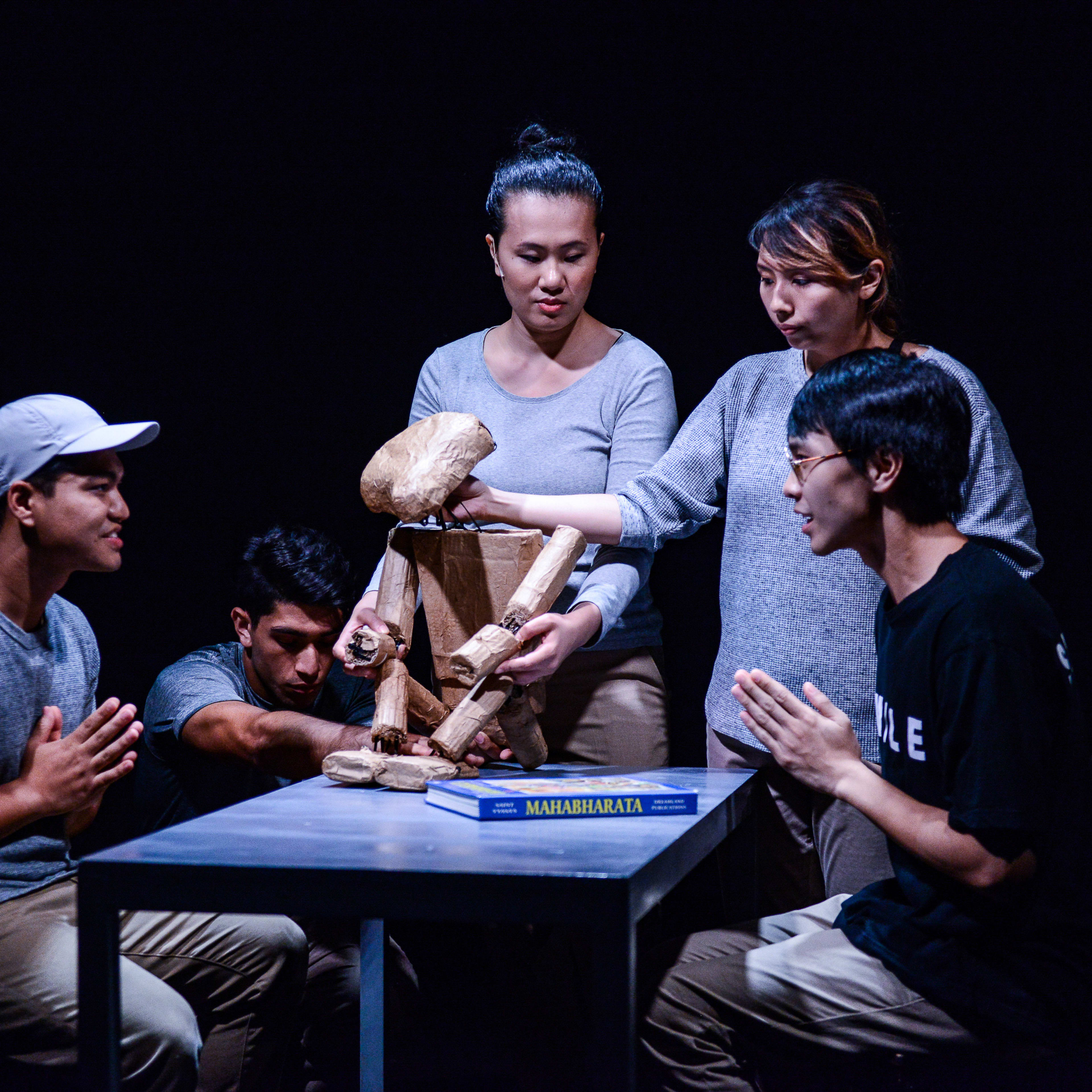 在天台製作（Rooftop Productions）的當代劇場作品《山下的證詞》（Testimony）中，四位演員在昏暗的排練室裡圍繞著一張桌子。兩位演員坐在兩側，雙手合十，神情虔敬；另外兩位則站在桌旁，細心操作著一個木製的骨架偶。這種演出氛圍呈現了香港當代舞台劇獨有的沉思與張力。其中一位表演者道出台詞：「當阿周那到達戰場的時候，他發現他的親戚在戰線對方，他不能戰鬥。」