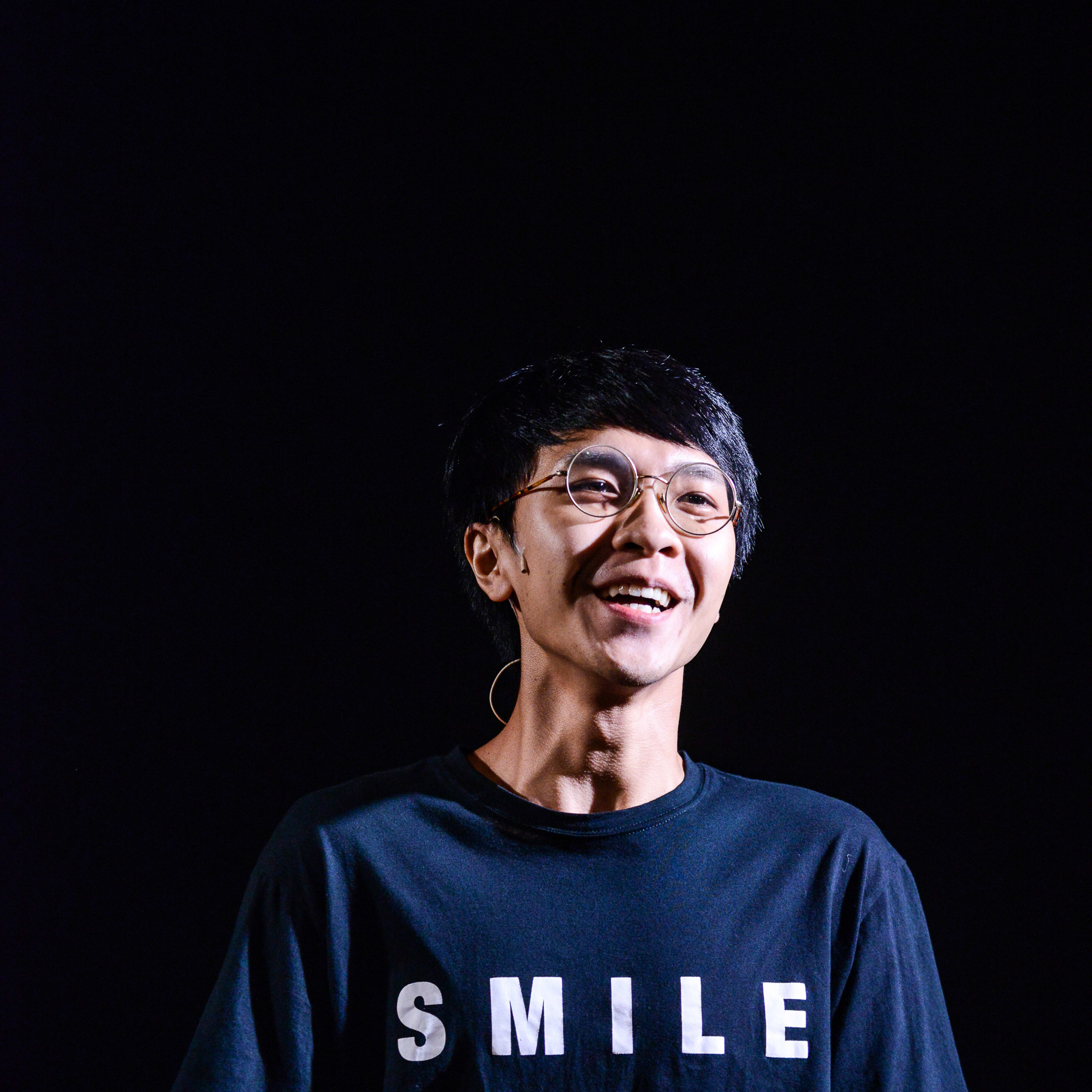 演員吳景隆站在簡約且幽暗的舞台上，聚光燈映照出他充滿感染力的笑容。他身穿一件印有「SMILE」字樣的黑色T恤，展現了香港當代劇場的極簡美學。背景投影出與劇目相關的文字，這張照片捕捉了天台製作（Rooftop Productions）劇目《山下的證詞》中的動人瞬間。《山下的證詞》