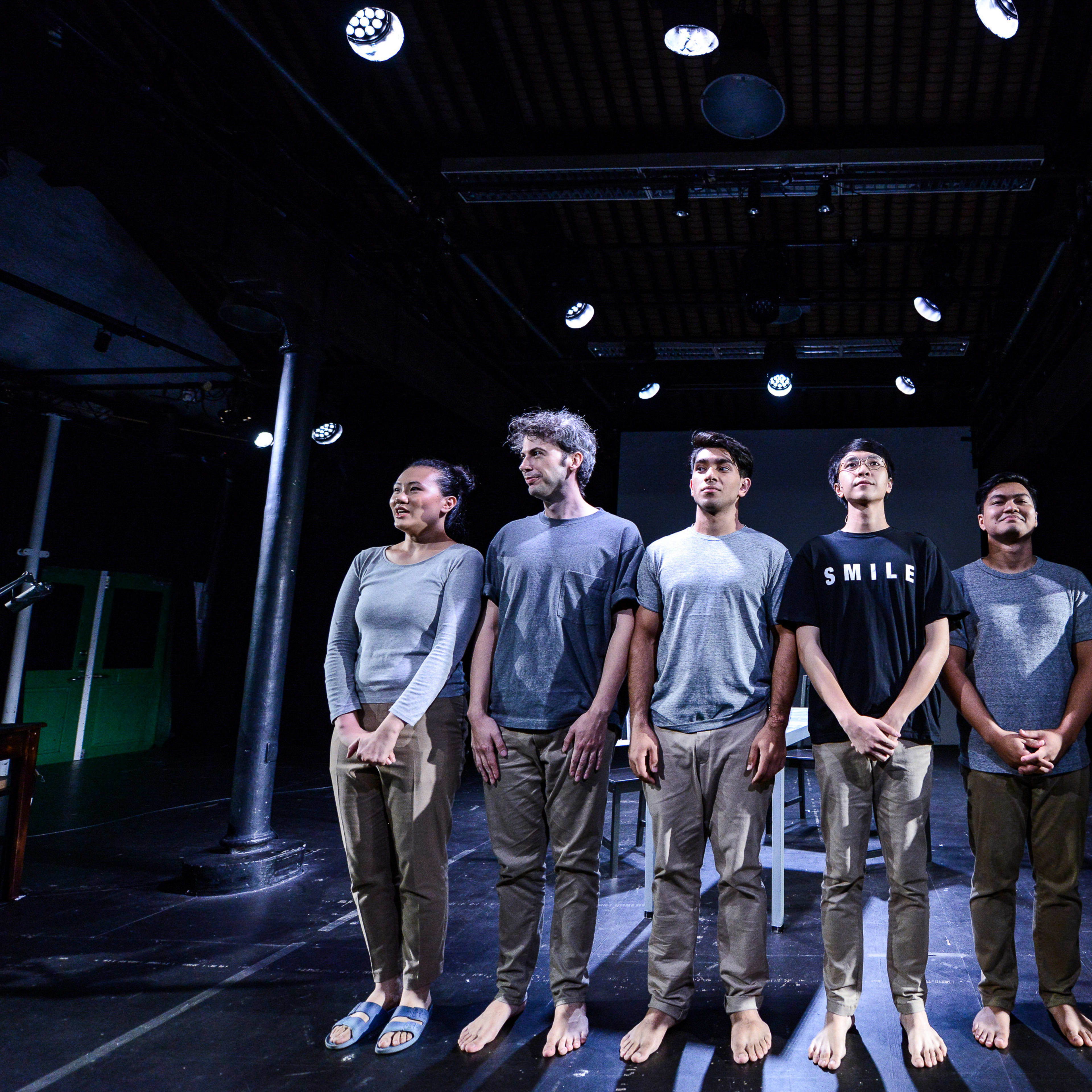 六位演員於天台製作（Rooftop Productions）的當代劇場作品《山下的證詞》舞台上並排站立。演員們穿著大地色系的休閒服裝，在昏暗的劇場燈光下齊齊仰望，神情莊嚴且深沉，呈現出香港當代實驗劇場獨有的情感張力與舞台美學。《山下的證詞》