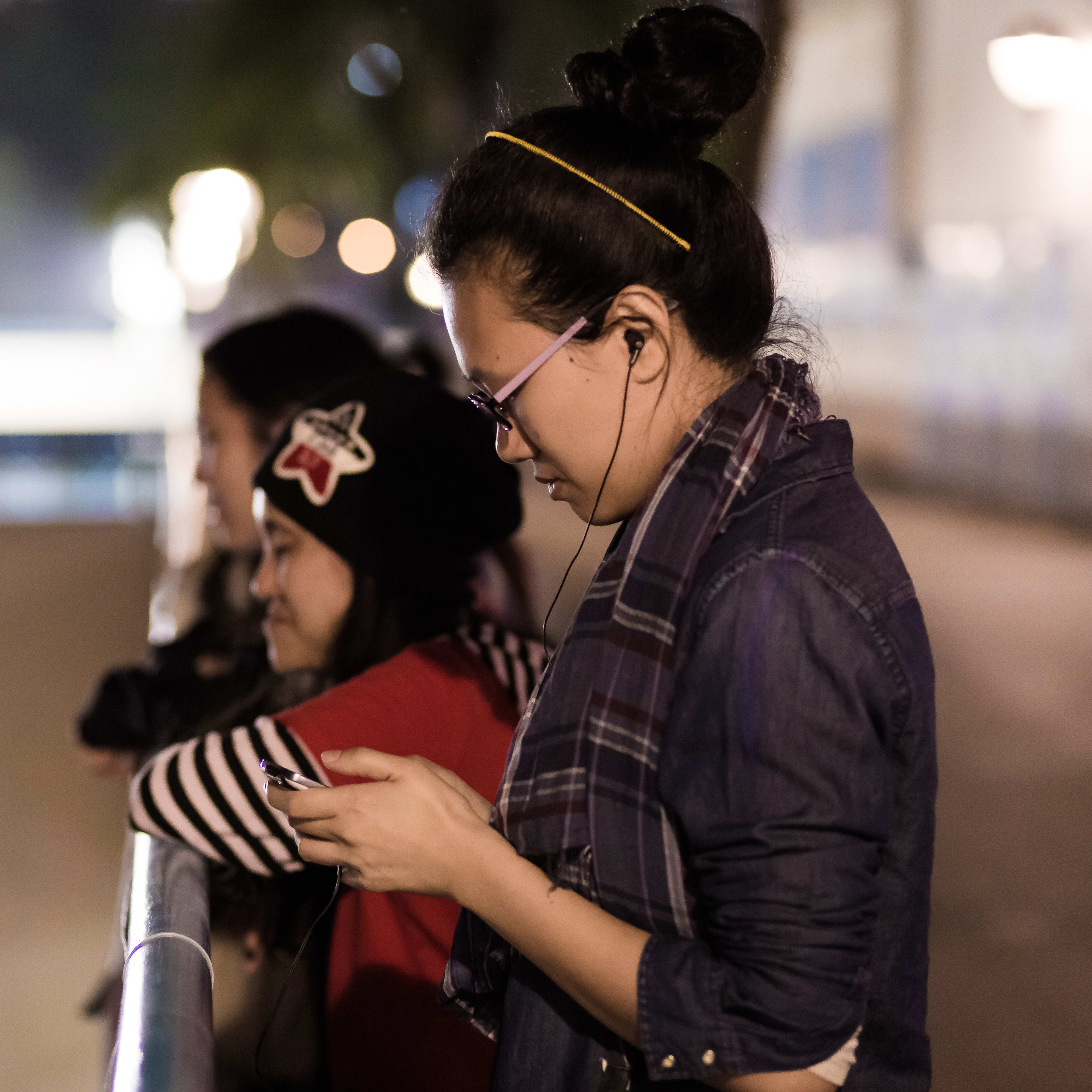 在天台製作（Rooftop Productions）當代劇場作品《行為淪喪》（The Beautiful Ones）的劇照中，三名女子在深夜的欄杆旁並肩而立，燈光昏暗且充滿都市氛圍。位於前景的李婉晶（Michelle Li）側身低頭，戴著眼鏡和耳機專注地注視手機螢幕；後方另一名頭戴星型圖案毛帽的演員則眺望城市夜景，呈現出該劇獨有的城市疏離感與親密氣息。《行為淪喪》
