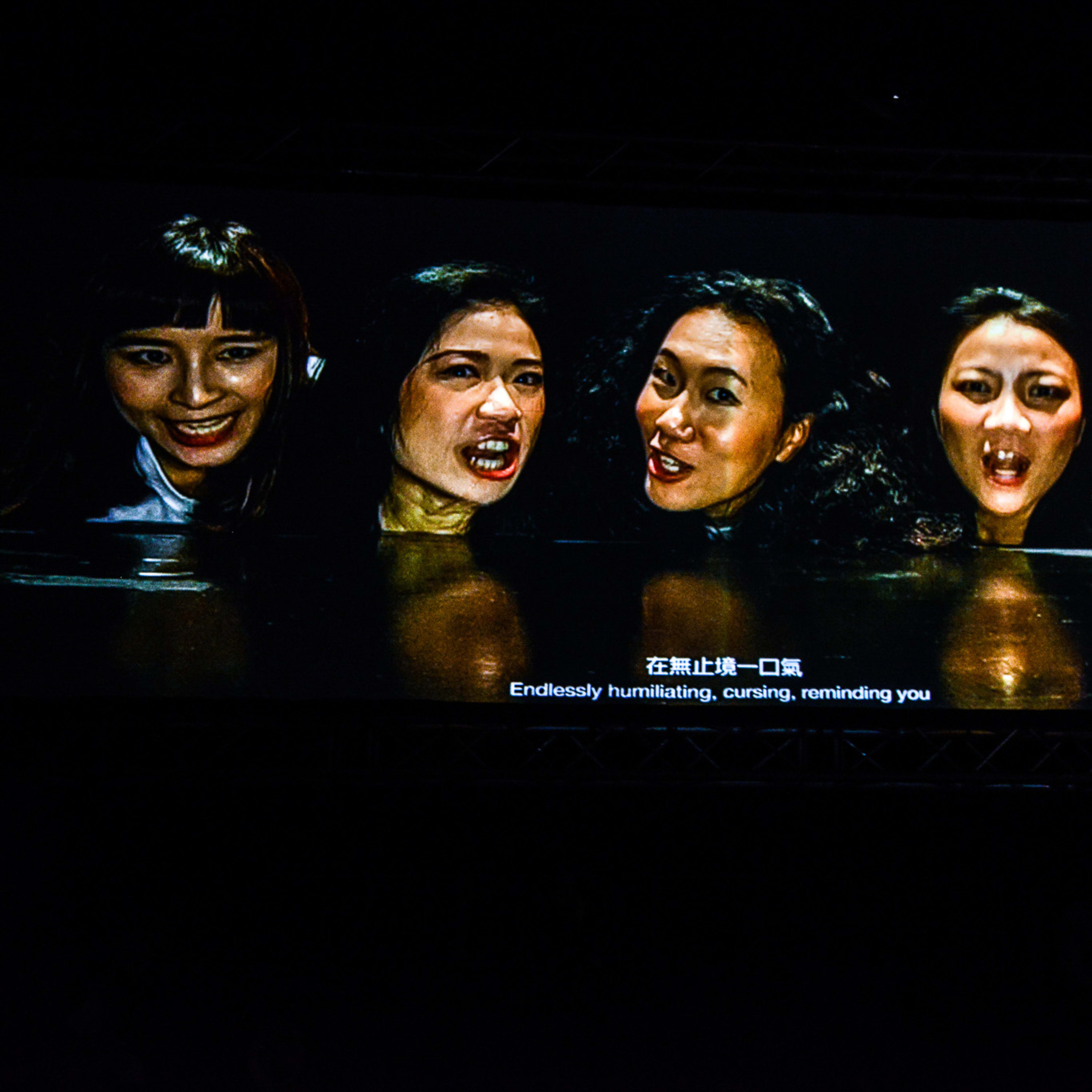 天台製作（Rooftop Productions）現代劇場作品《復仇變奏曲》（The Furies Variations）的劇照，燈光昏暗強烈，五位演員一字排開，神情猙獰激動，展現出香港實驗劇場特有的強烈張力。前景中一位表演者的剪影與舞台中央的陣容形成呼應。角色們在台上唸出：「在無止境一口氣侮辱詛咒提醒你犯下弒母的血仇。」