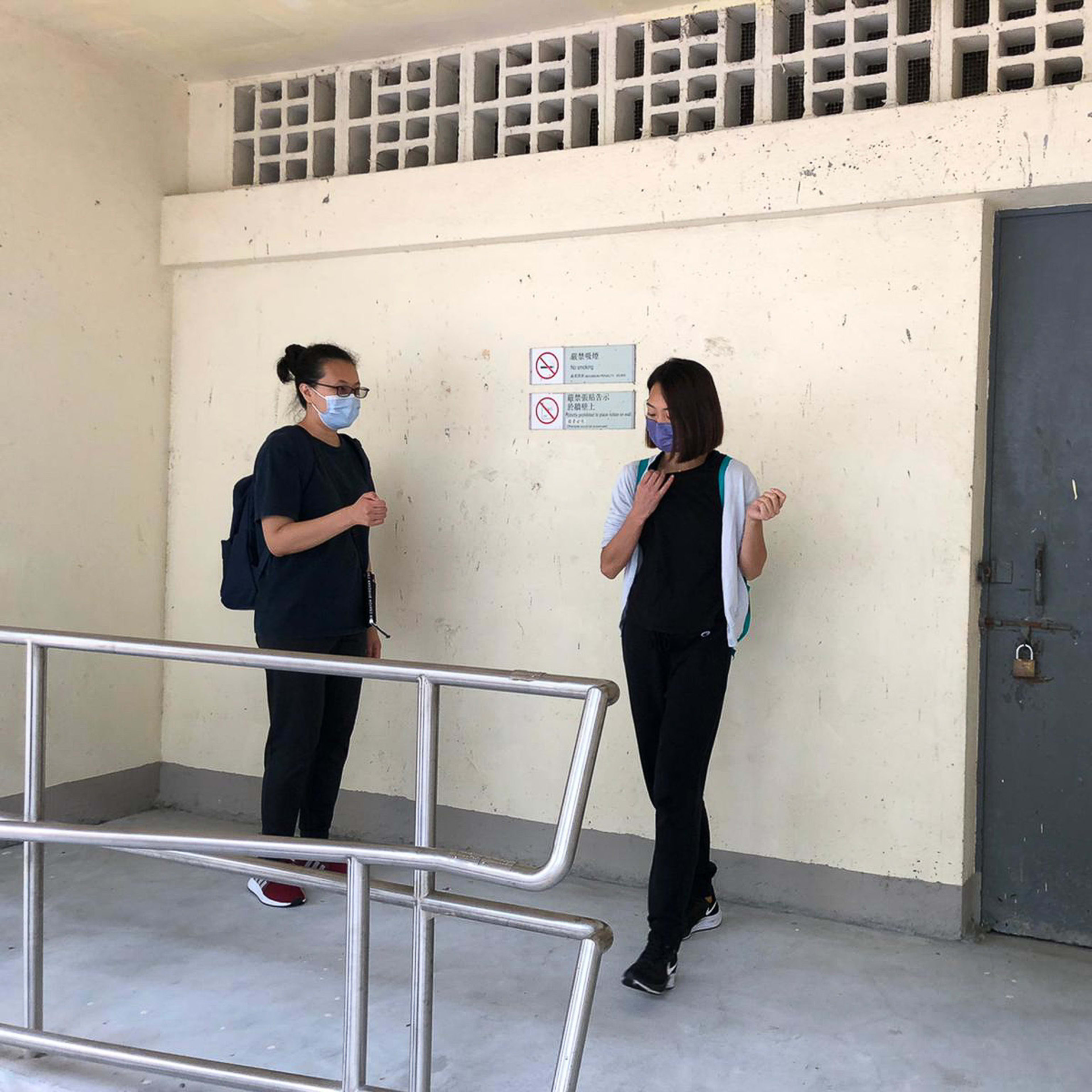 兩位演員於香港當代劇場製作《舞照跳》的排練場地對話，背景是簡約樸實的混凝土走廊。穿著黑色短袖上衣的周芳站在左側，李婉晶則穿著白色外搭與黑色上衣，正走向場地中央。此影像捕捉了排練期間的靜謐時刻，展現《舞照跳》對藝術堅持與現實生存的深刻探討。其中一位演員在對話中思索道：「現實意義。」