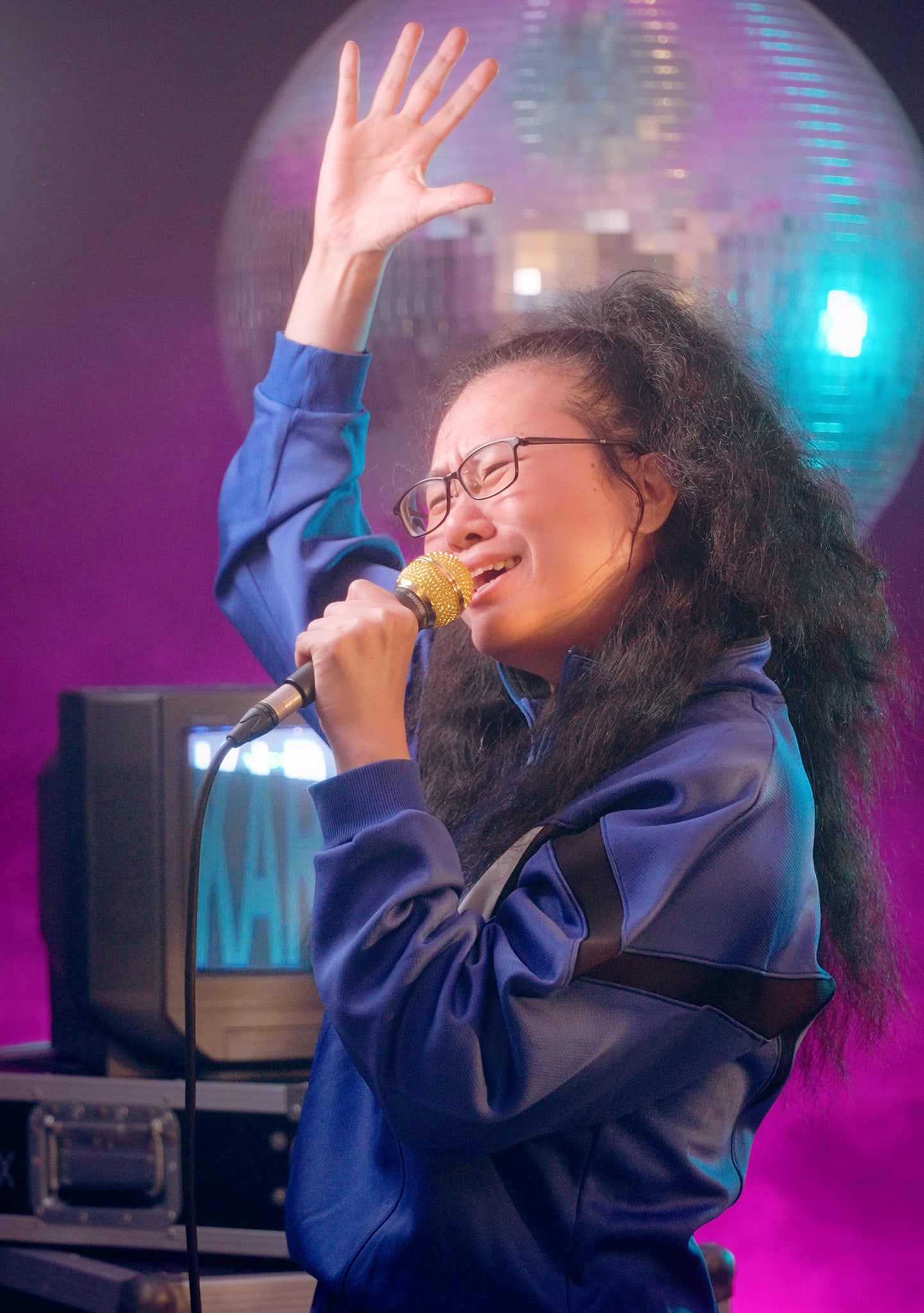 這張充滿戲劇張力的特寫照片捕捉了表演者李婉晶在天台製作當代劇場作品《卡啦OK》中的演出瞬間。李婉晶身穿藍色運動套裝，戴著眼鏡，在舞台燈光的照耀下，深情地對著金色麥克風演唱，左手高高舉起，充滿感染力。背景中的鏡面球和復古電視機為這場西九文化區（WestK）的演出營造出獨特的懷舊氛圍。《卡啦OK》KV。