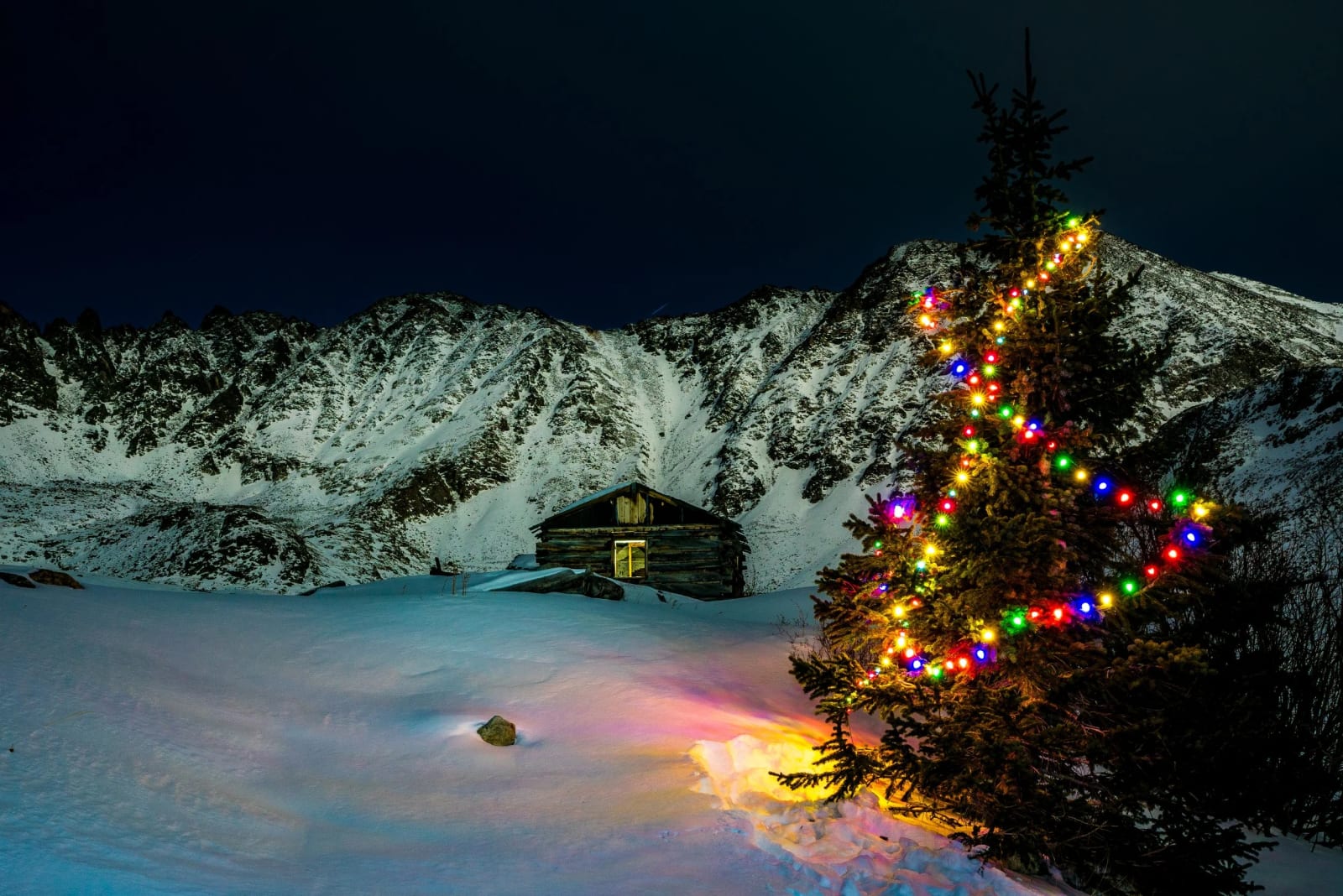 December at the Boston Mine — Mayflower Gulch, Colorado. A lit Christmas tree standing beside the old Boston Mine cabin under a clean night sky at Mayflower…