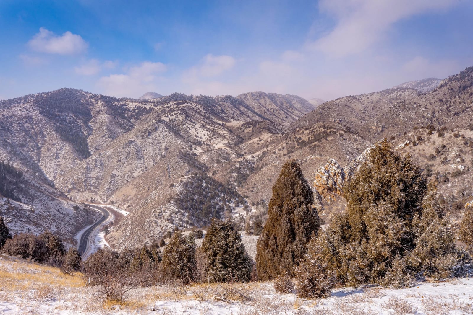 From Up Here the Traffic Is a Rumor — Clear creek canyon. A Colorado highway threading the canyon floor far below under a fresh dusting of snow, juniper and…