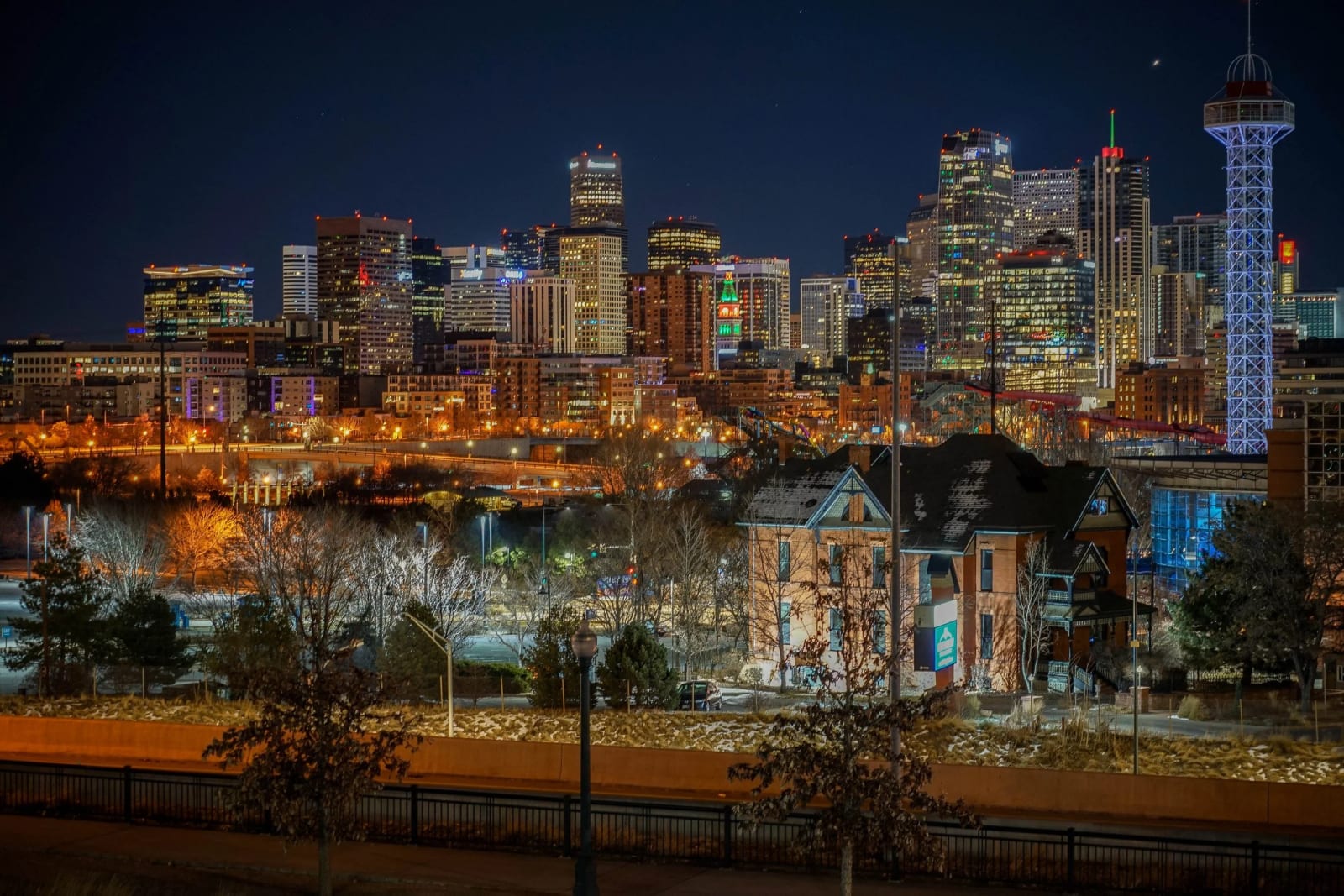 The House That Watched It All Arrive — Denver, Colorado. Downtown Denver glowing behind one of the old brick survivors on the west side, holiday lights…