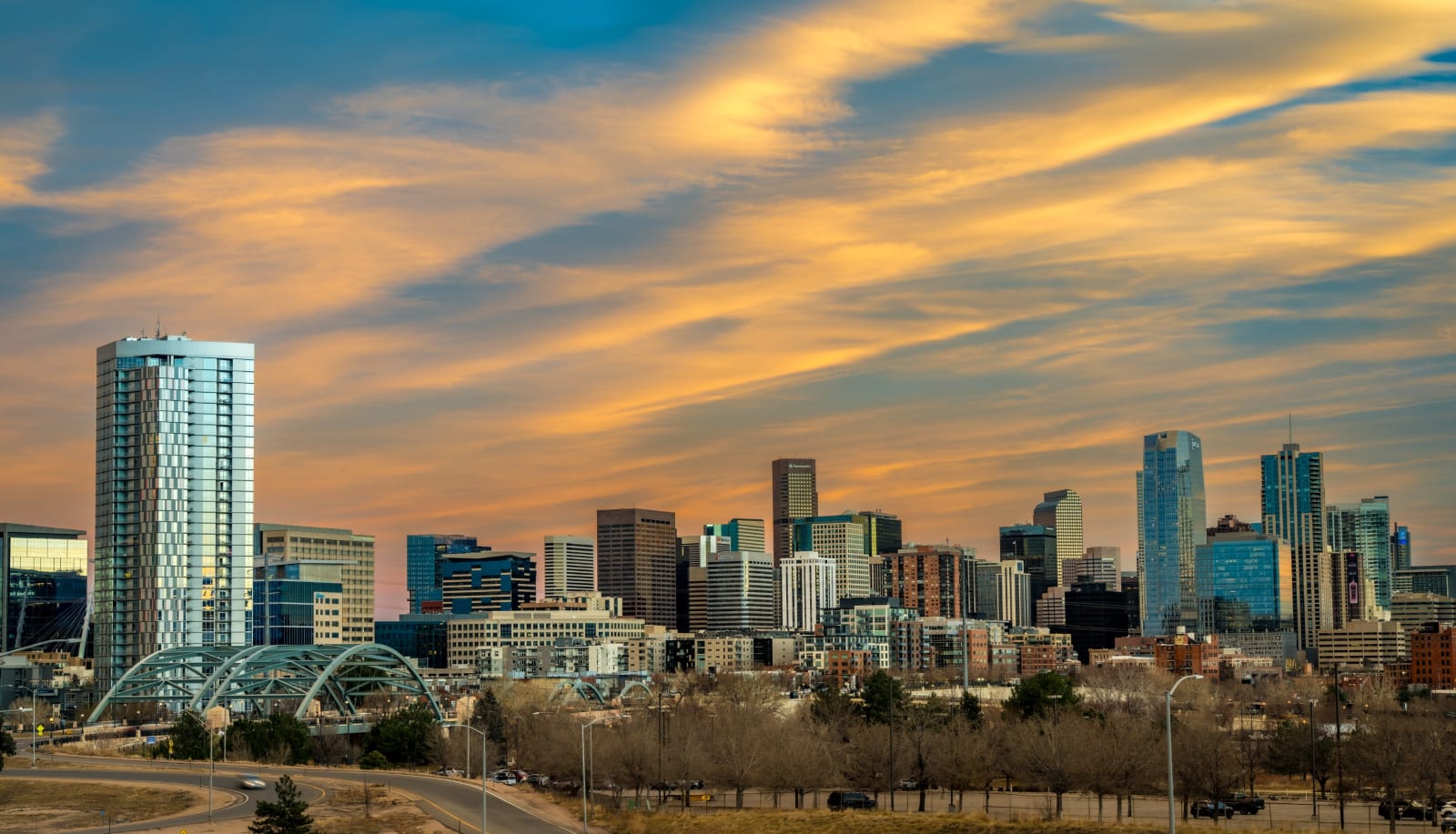 Winter Light on the Skyline — Denver, Colorado. The Denver skyline stretched out under a full sky of gold-lit cirrus, bare trees holding the foreground and…