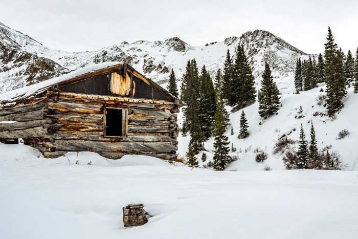 They Built for Keeps — Mayflower Gulch, Colorado. A hand-hewn log cabin with a sagging roof still shrugging off the drifts at eleven thousand feet in…