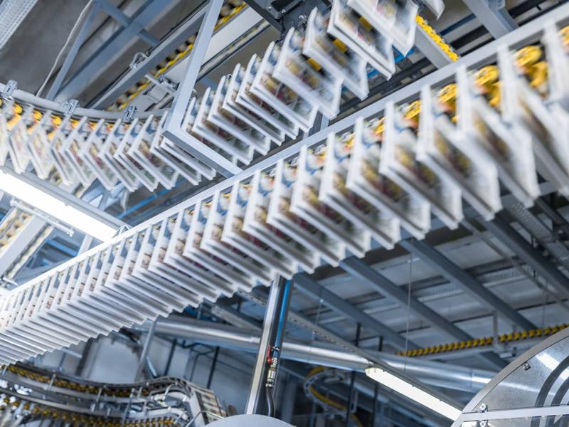 The image shows a production line being transported by a conveyor belt system within a printing press facility. 
