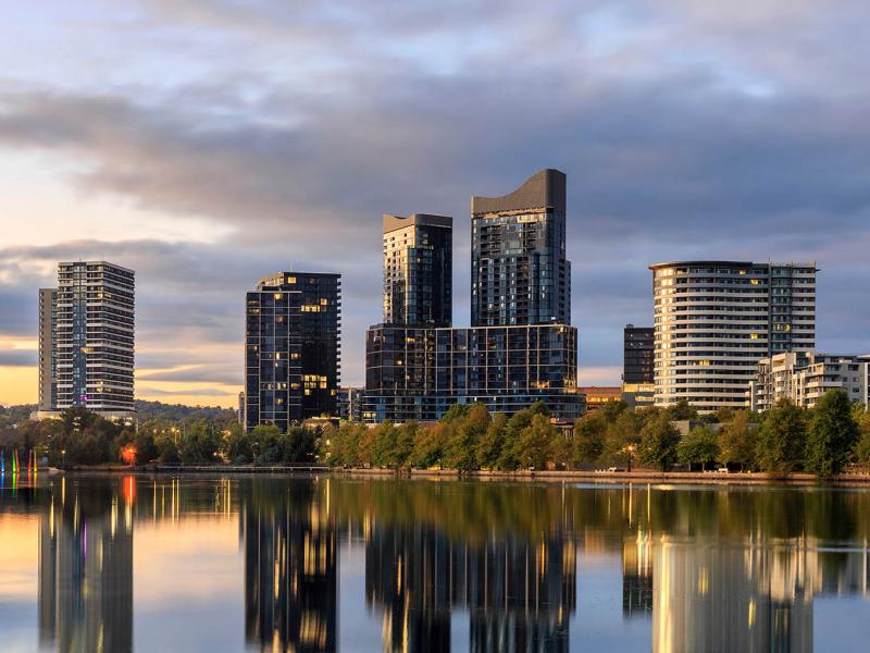 The image displays the cityscape of Canberra, Australia, specifically featuring buildings reflected in Lake Burley Griffin. 