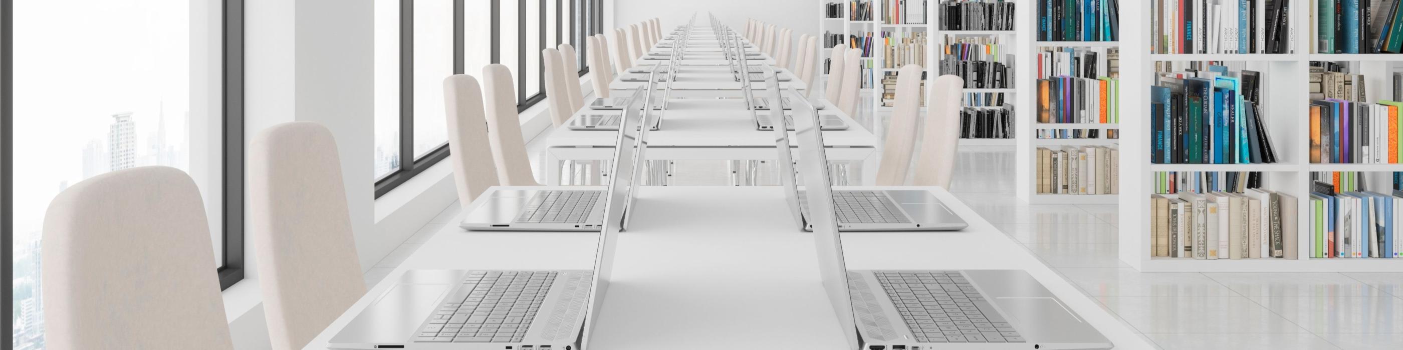 A spacious, modern library study area with long white tables lined with open laptops and beige chairs. 