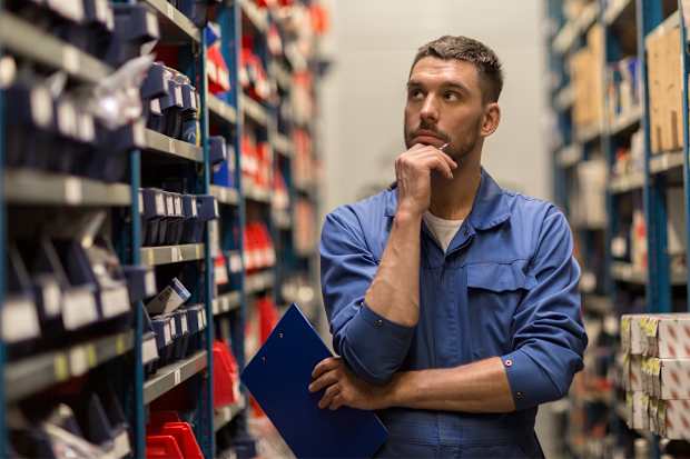 Warehouse worker holding a clipboard while inspecting stocked shelves and considering inventory levels.