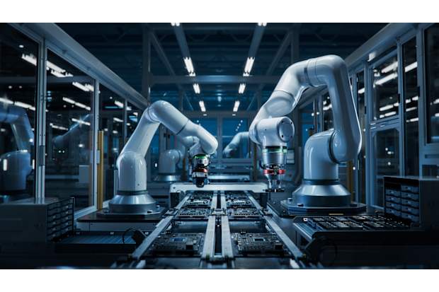 Two robotic arms working over electronic circuit boards on an automated manufacturing line inside a modern, enclosed factory.