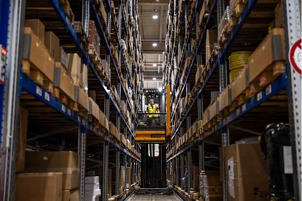 A warehouse forklift operator in a hi-vis vest driving a forklift through two large, narrow shelving units full of stock.