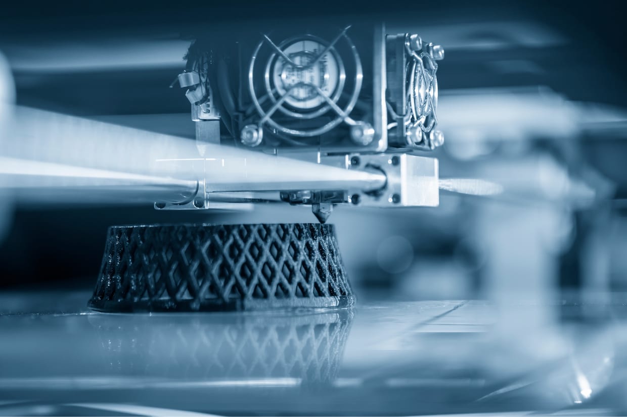 Close-up of a 3D printer in operation, depositing material to create a dark lattice-patterned object on the print bed, with the printer's extruder and cooling fans visible above.