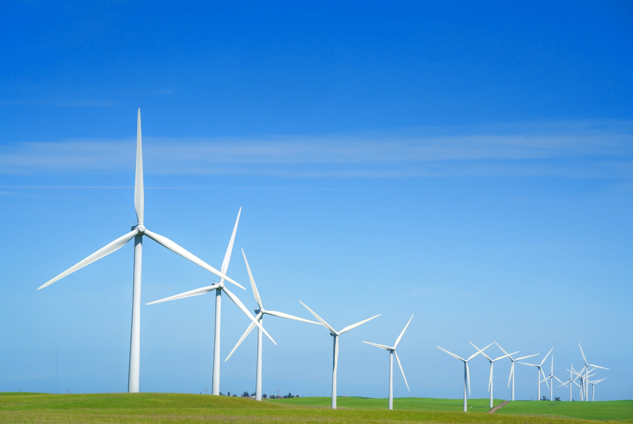 A line of operational wind turbines in a green field and blue sky generating power.
