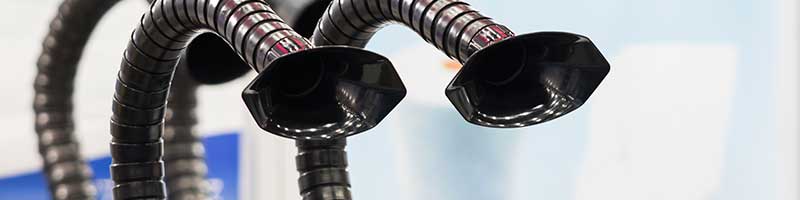 Close-up of two flexible fume extractor arms with black hood nozzles positioned to capture airborne fumes in a workspace.