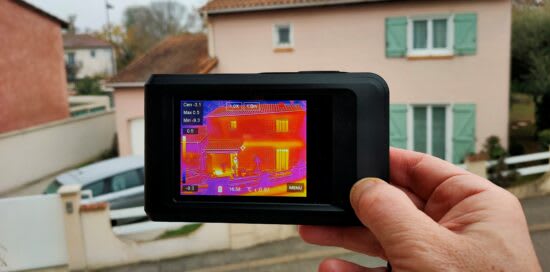 A handheld thermal imaging camera displaying a heat map of a two-story residential house.