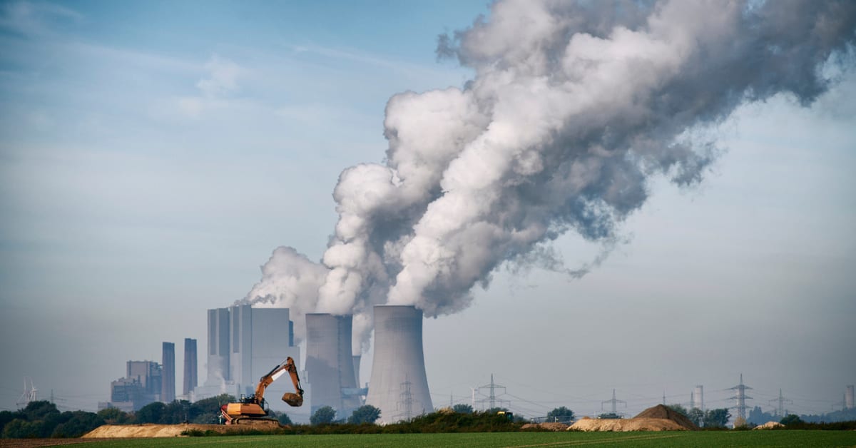 A distant shot of a fossil fuel power plant exhausting dark grey fumes from its cooling towers.