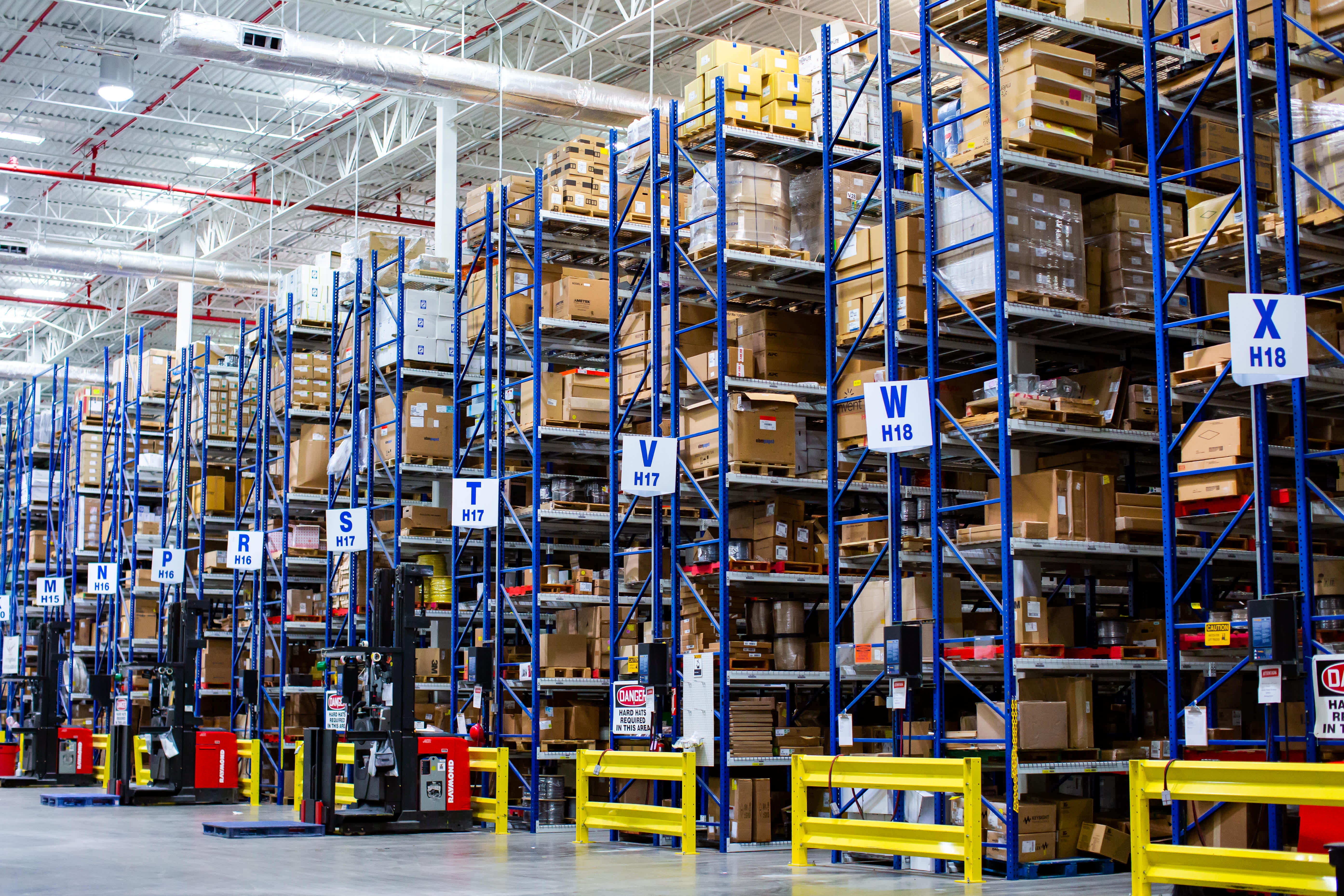 Large warehouse interior with tall blue pallet racks filled with cardboard boxes and packaged goods, labeled storage sections, forklifts, and yellow safety barriers along the aisles.