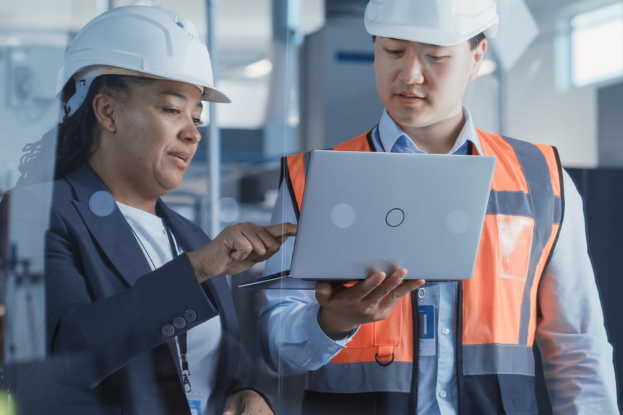 2 engineers discussing over a laptop, wearing proper PPE in hard hats and an orange hi-vis.