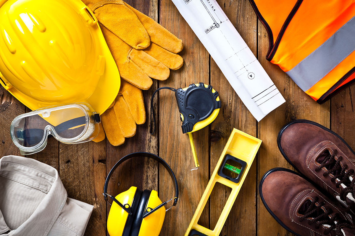 A collection of PPE equipment laid out, including a yellow hard hat, protective gloves, safety goggles, ear protection, a measuring tape, a spirit level, a hi vis vest, and work boots.