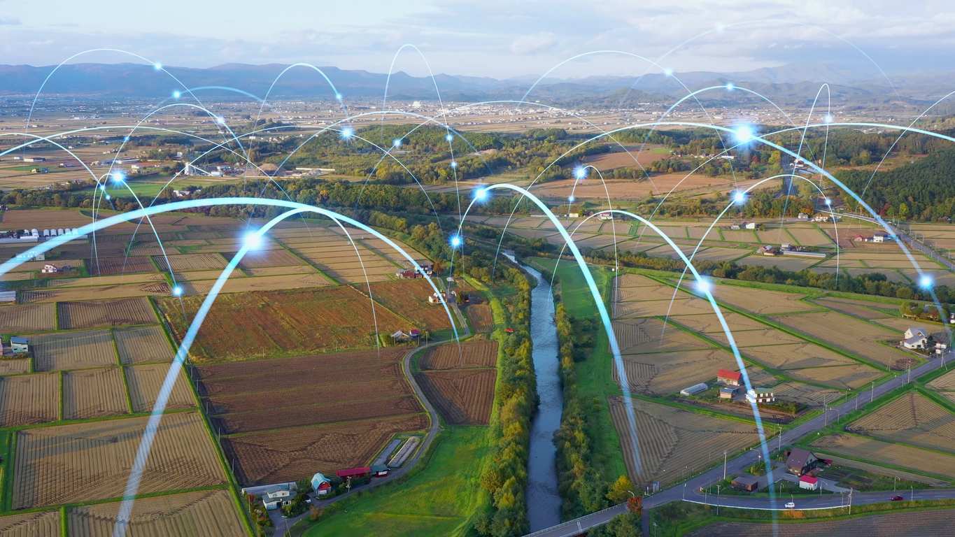 Aerial view of rural farmland with connected digital network lines linking fields, roads, and homes, illustrating smart village technology and connected agriculture infrastructure.