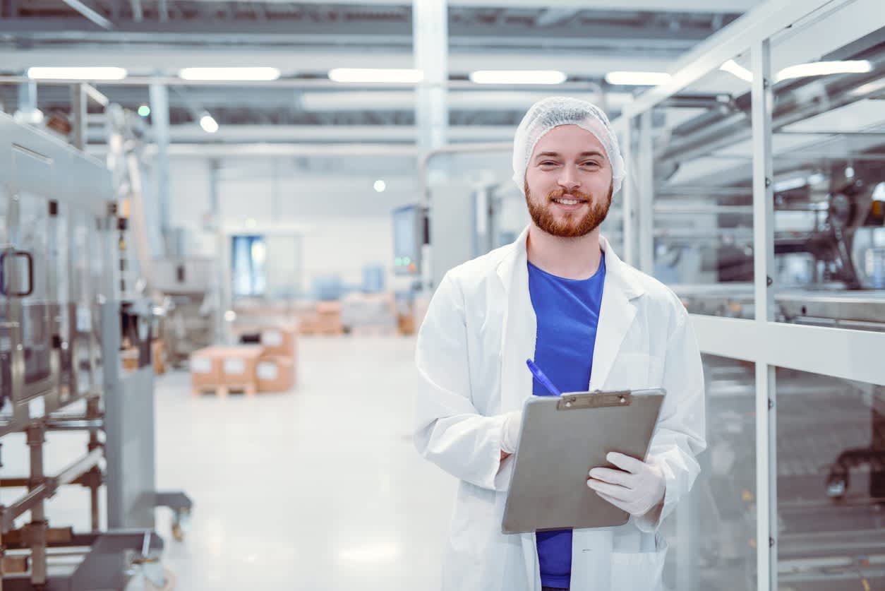 A man in a food processing centre surrounded by ovens and conveyor belts dressed in PPE, holding a clipboard and pen.