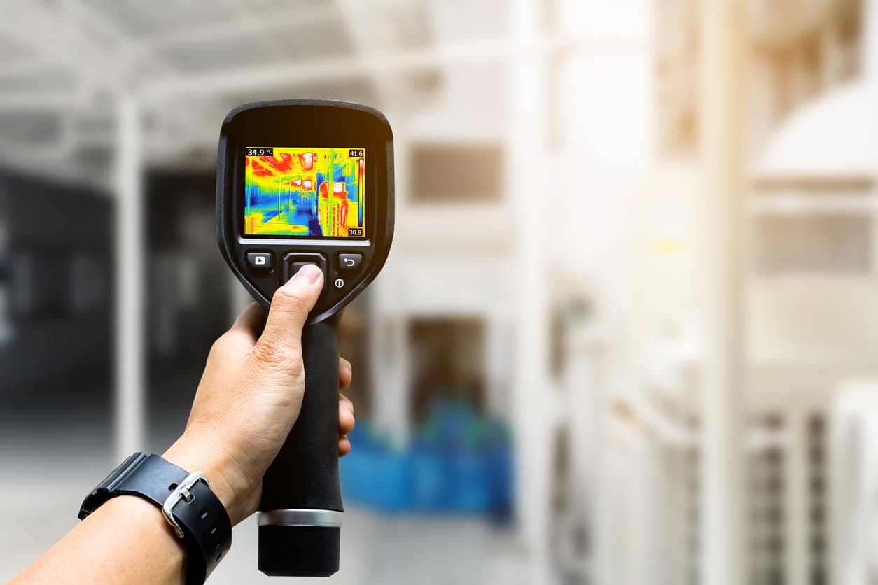 A person pointing a thermal imaging camera at machines, with the camera displaying heat signatures emitting from the machines.