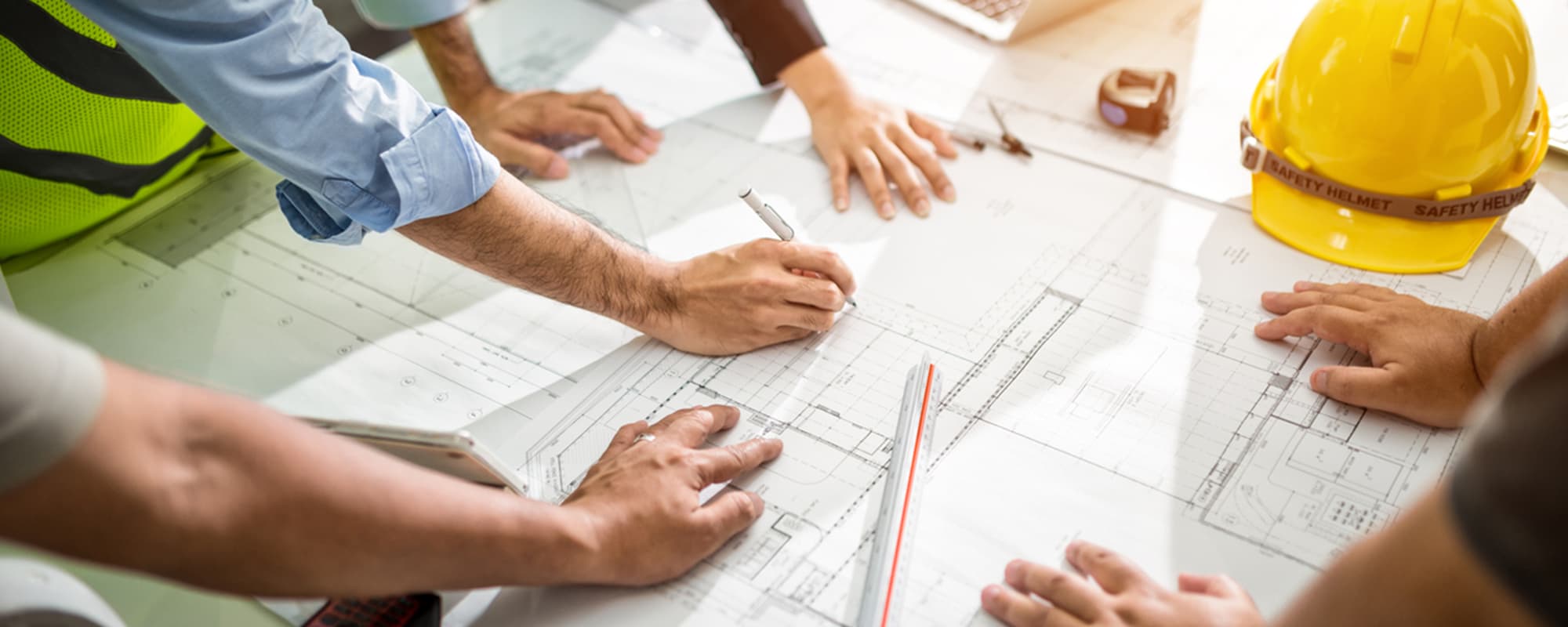 Engineers surrounding a table, inspecting blueprints of a building with measuring tools and a yellow hard hat on top of the surface.