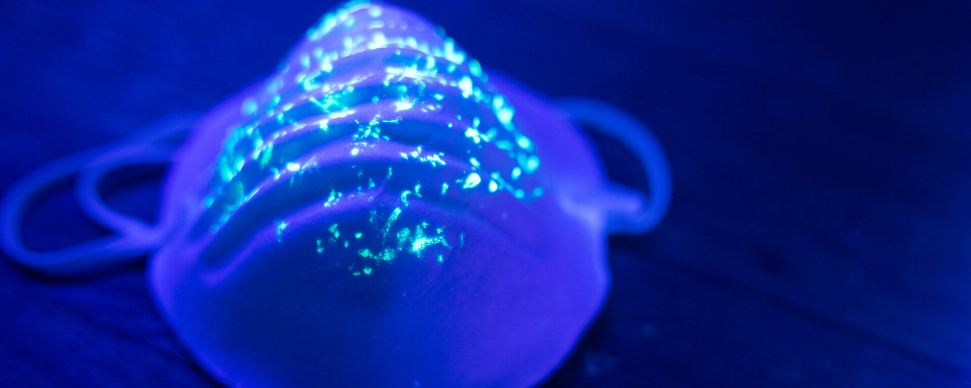 Medical face mask under ultraviolet (UVC/UV) lighting showing glowing fluorescent particles and contamination spots across the surface.